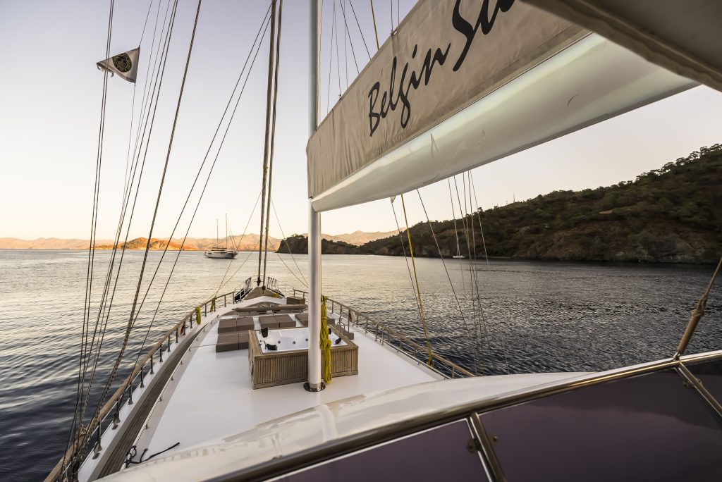 View from the deck of a sailboat at sunset, with calm water and hills in the background. The boat’s seating area is visible, and distant sailboats can be seen near the shoreline.