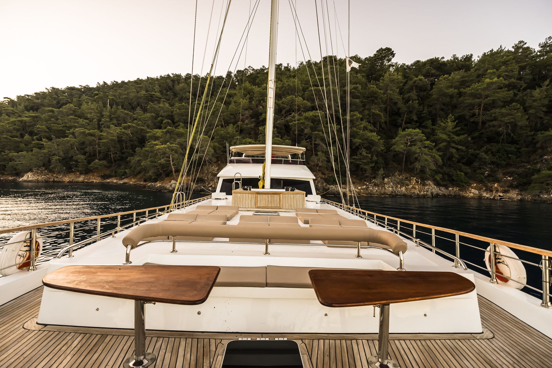View from the deck of a yacht facing forward, with seating and two wooden tables visible. The yacht is anchored near a forested shoreline with calm water, under a clear sky at sunset.