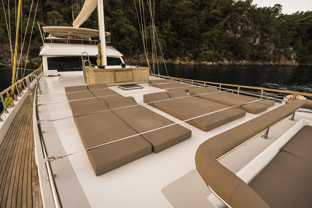 A luxury yacht deck with several brown sunbeds arranged neatly, surrounded by railings, with forested cliffs and calm water in the background.