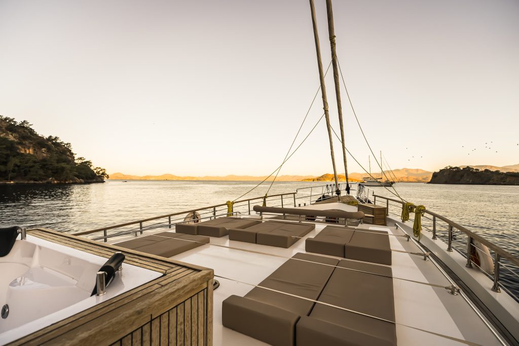 View from the deck of a yacht at sunset, showing cushioned lounge areas, a hot tub, calm sea, distant mountains, and small islands under a clear sky.