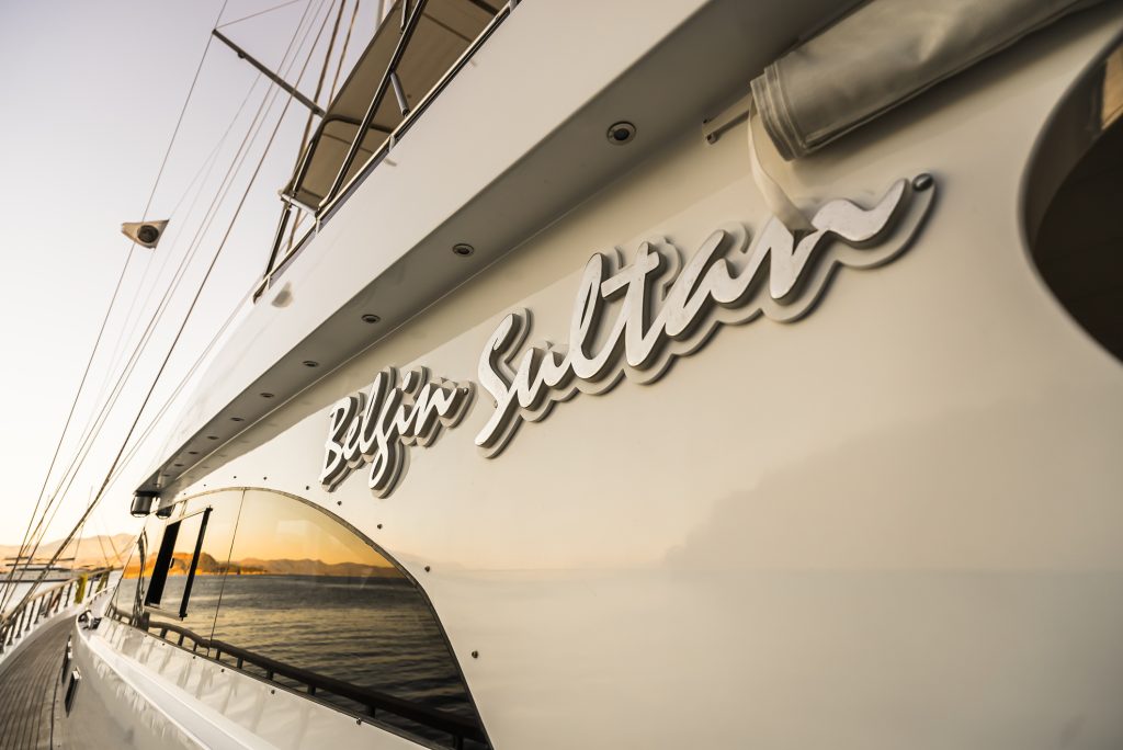 Close-up of a white yacht named BILGIN SULTAN, with the name displayed in silver letters on the side, calm water and coastline reflected in the yachts windows at sunset.