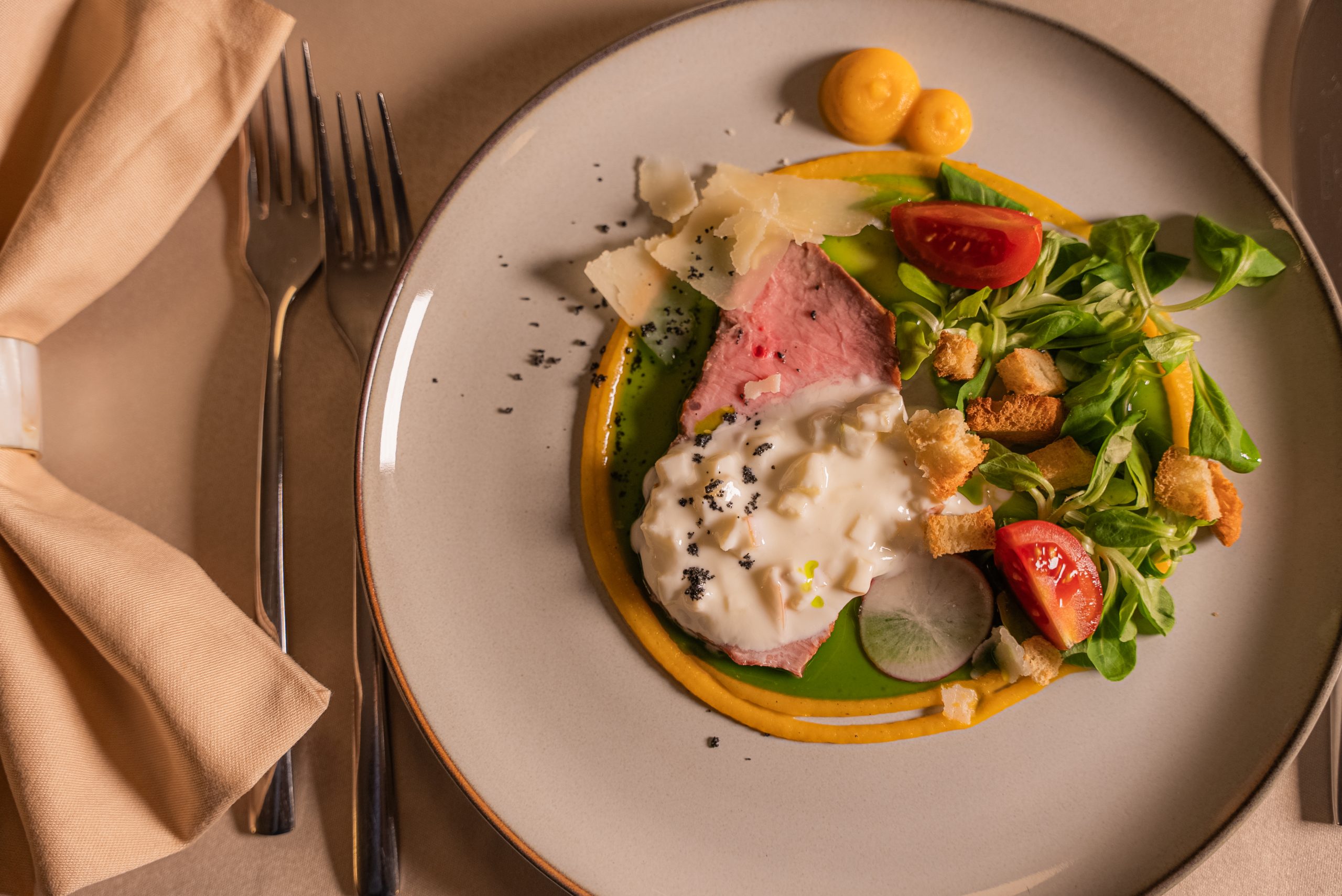 A plated dish featuring sliced roast beef topped with a creamy white sauce, garnished with greens, cherry tomatoes, croutons, radish, Parmesan, and yellow sauce swirls on a beige plate with a fork, knife, and napkin beside it.