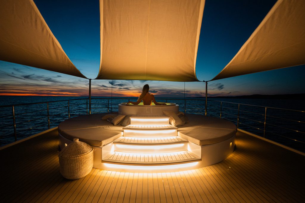 A person sits in a round, illuminated hot tub on a yacht deck at sunset, under a canopy, looking out over calm ocean waters with a vibrant, colorful sky in the background.