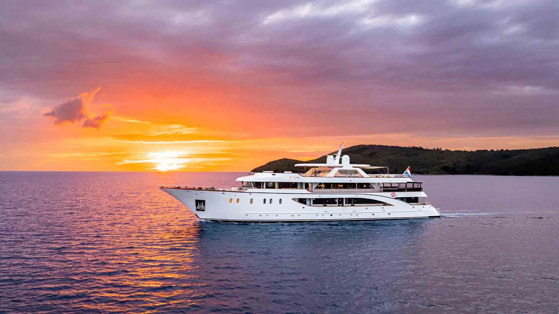 CRISTAL A large white luxury yacht sails on calm blue water at sunset, with a vibrant orange sky and clouds. An island with green hills is visible in the background.
