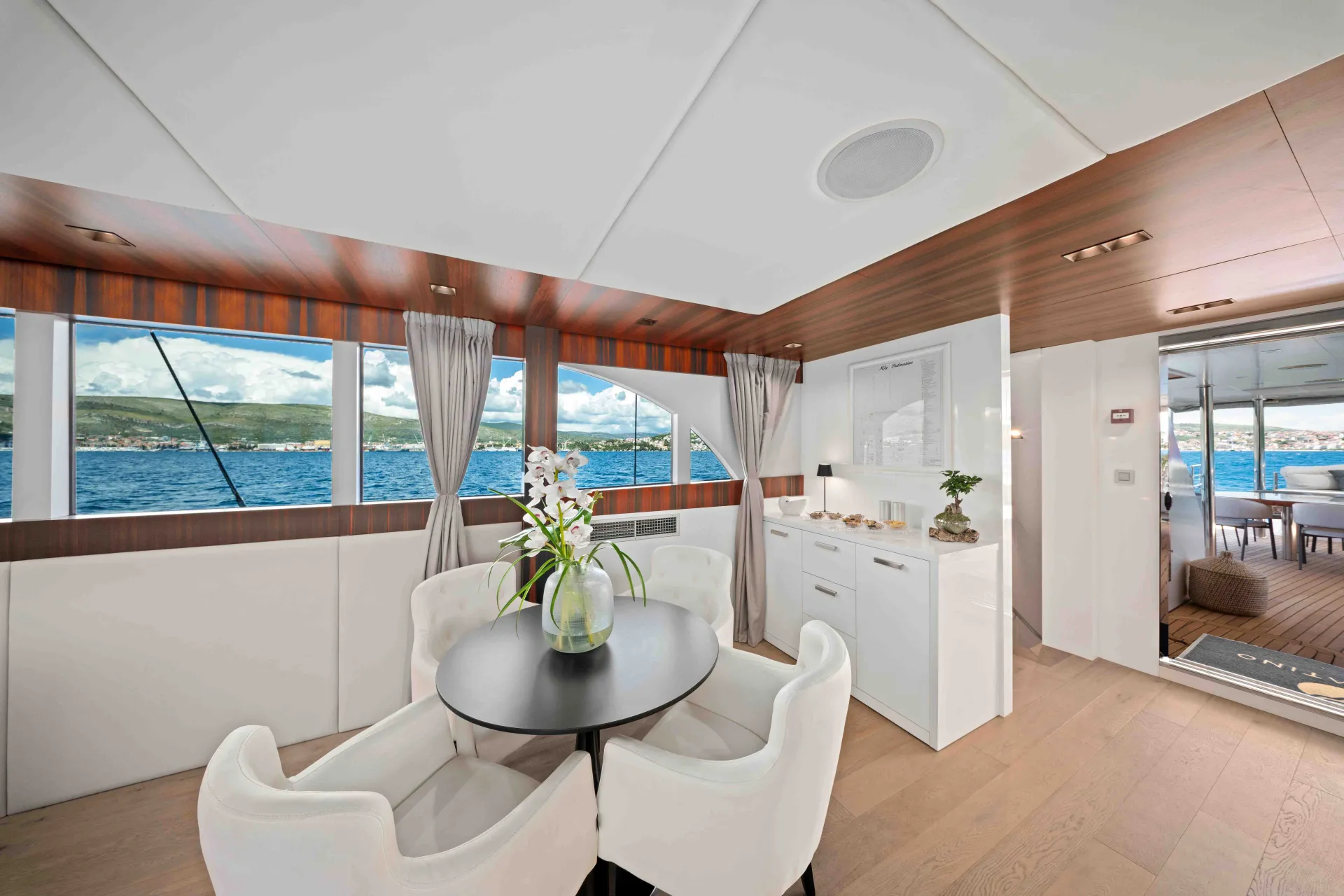 A modern yacht interior with a round dining table, white chairs, a vase with flowers, large windows offering ocean views, and a kitchenette. Bright, airy, and decorated with light wood and neutral tones.
