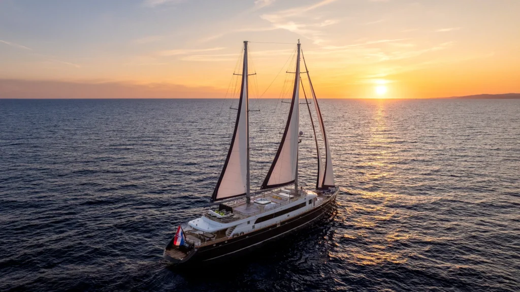 A large sailboat with two masts sails on calm water at sunset, with the sky painted orange and yellow as the sun nears the horizon. The sea stretches endlessly, reflecting the warm colors of the sky.
