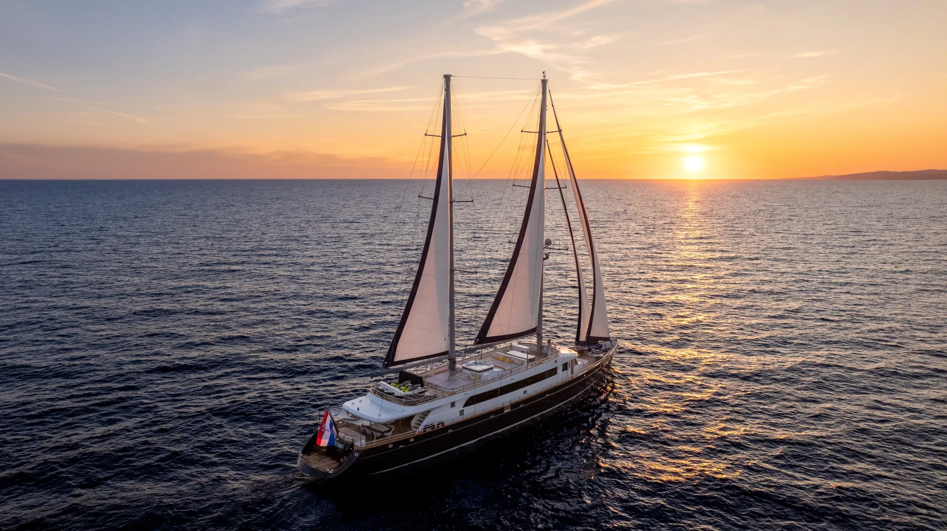 A large sailboat with two masts sails on calm water at sunset, with the sky painted orange and yellow as the sun nears the horizon. The sea stretches endlessly, reflecting the warm colors of the sky.