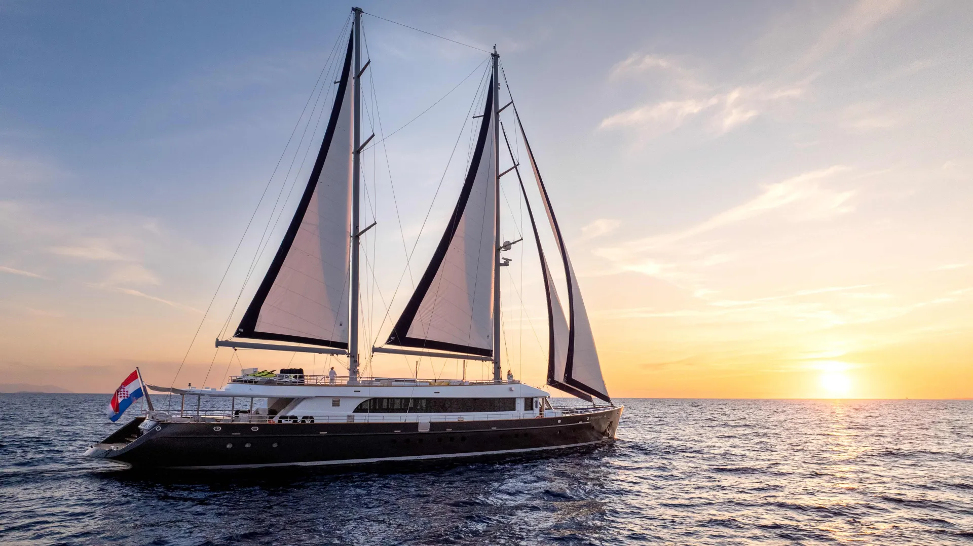 A large, modern sailing yacht with two masts glides on calm ocean waters at sunset, with the sun low on the horizon and a colorful sky in the background. A flag waves at the stern of the yacht.