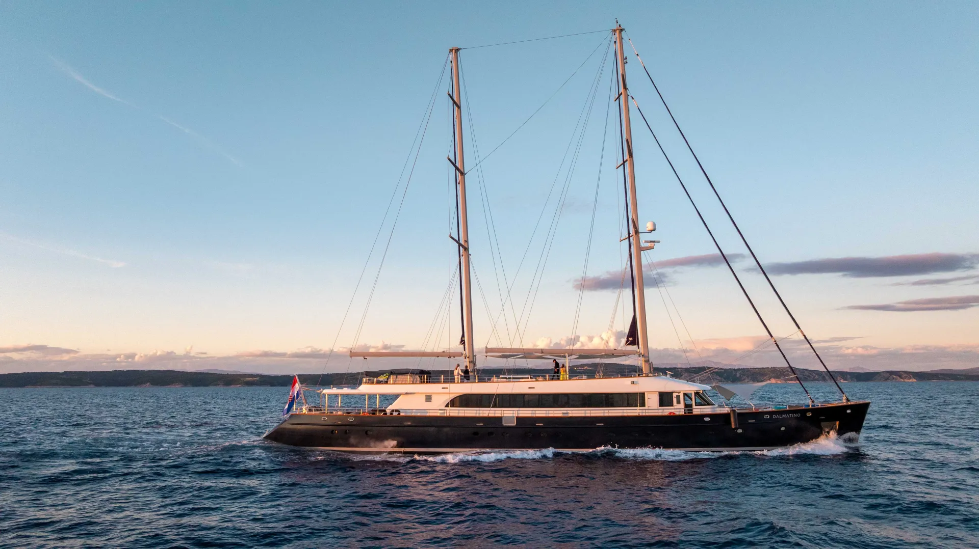 A large, modern sailing yacht with two tall masts cruises on calm blue water at sunset, with a hilly coastline and pastel sky in the background.