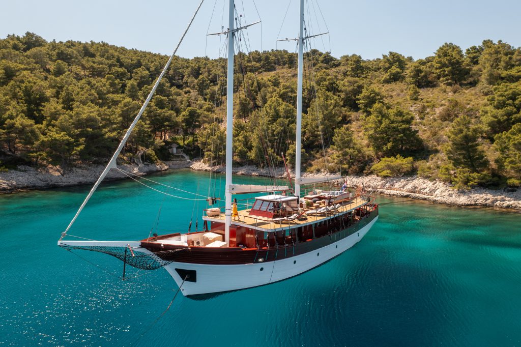 ROMANCA A large wooden sailboat is anchored in a calm, turquoise bay surrounded by rocky shores and dense green pine trees under a clear blue sky.