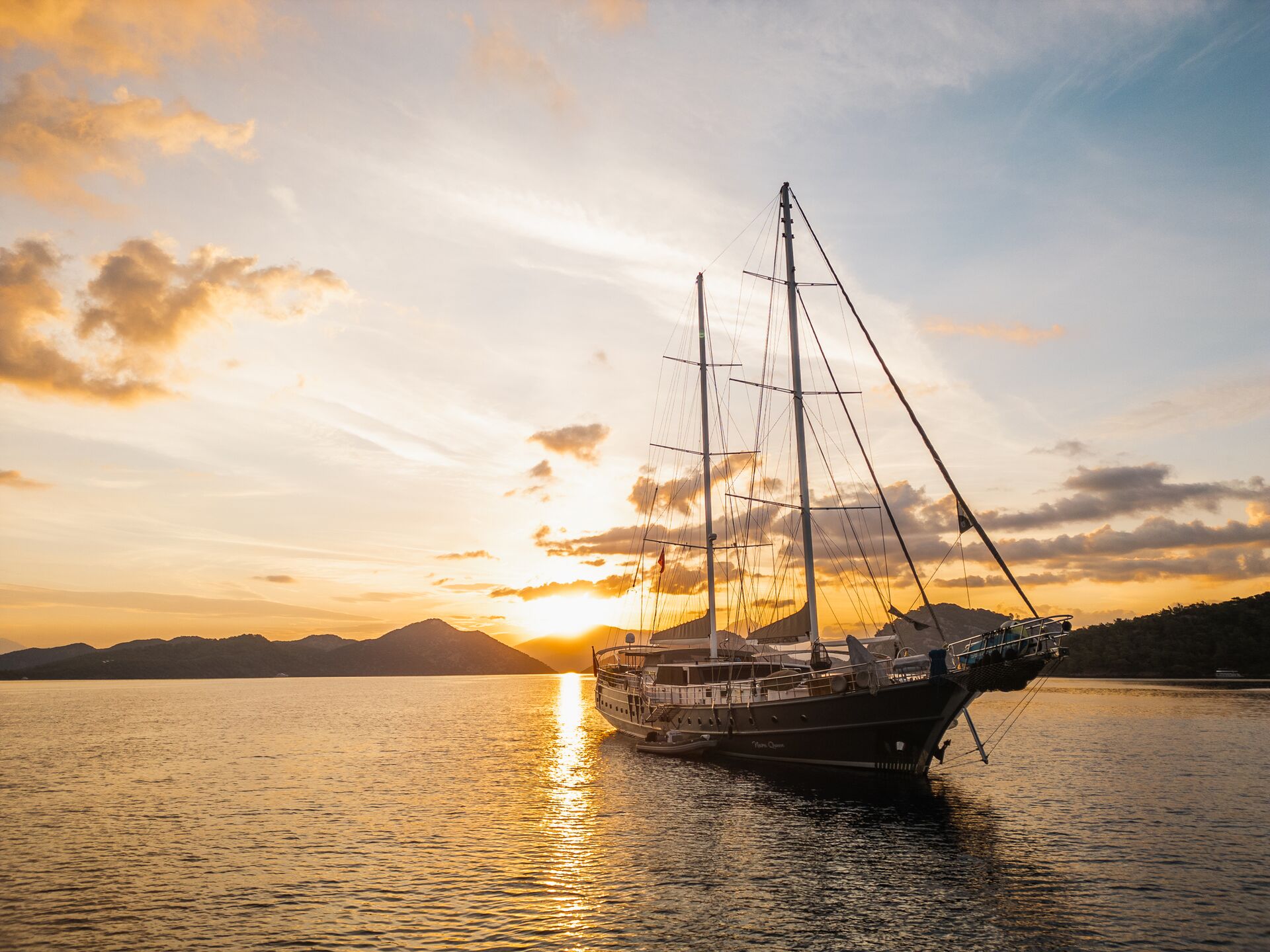 A sailboat with tall masts floats on calm water at sunset, with golden sunlight reflecting off the sea and silhouetted hills in the background under a partly cloudy sky.