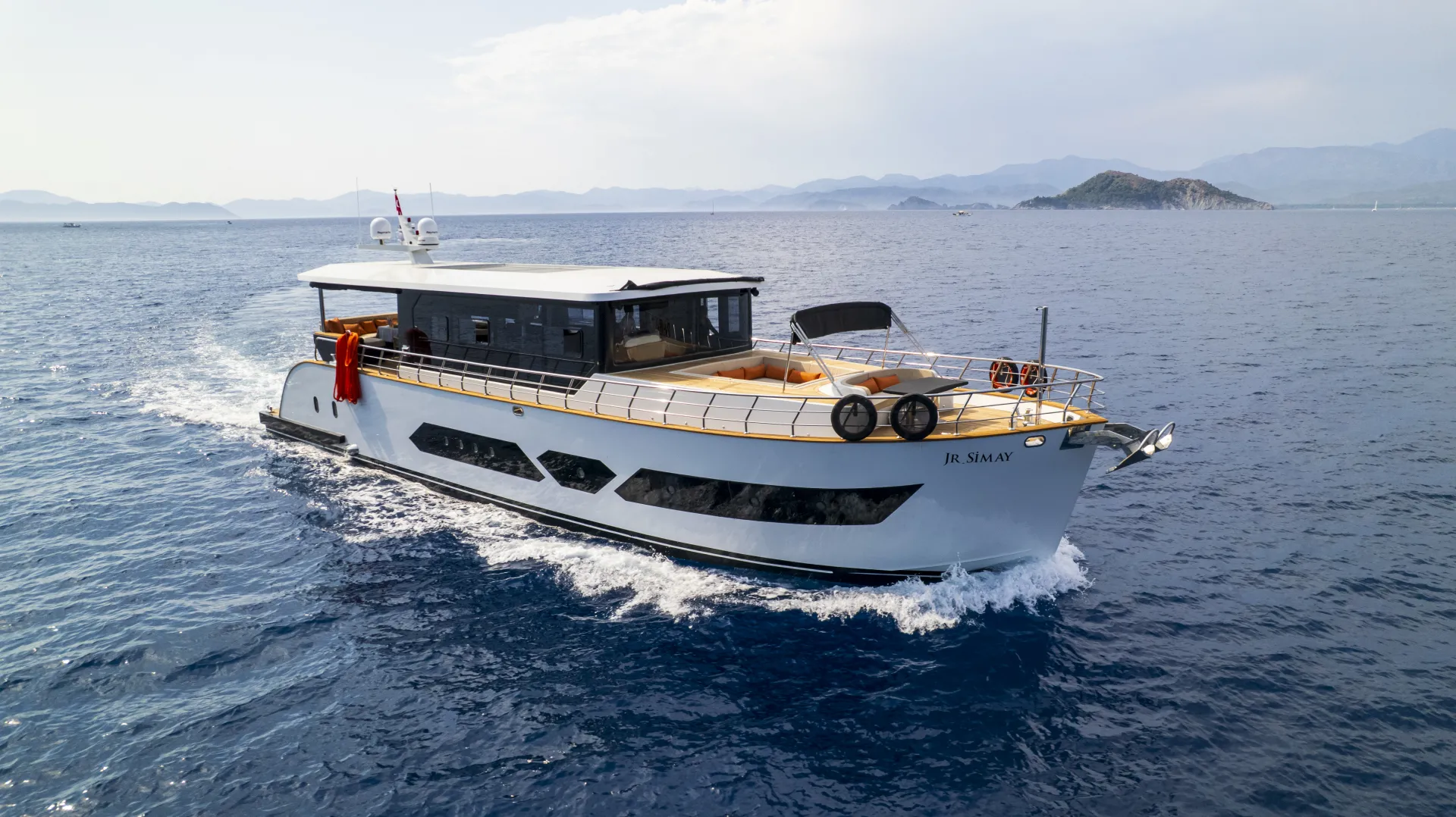 JUNIOR SIMAY A modern white yacht cruises through calm blue ocean waters near a distant coastline with mountains and an island visible under a partly cloudy sky.