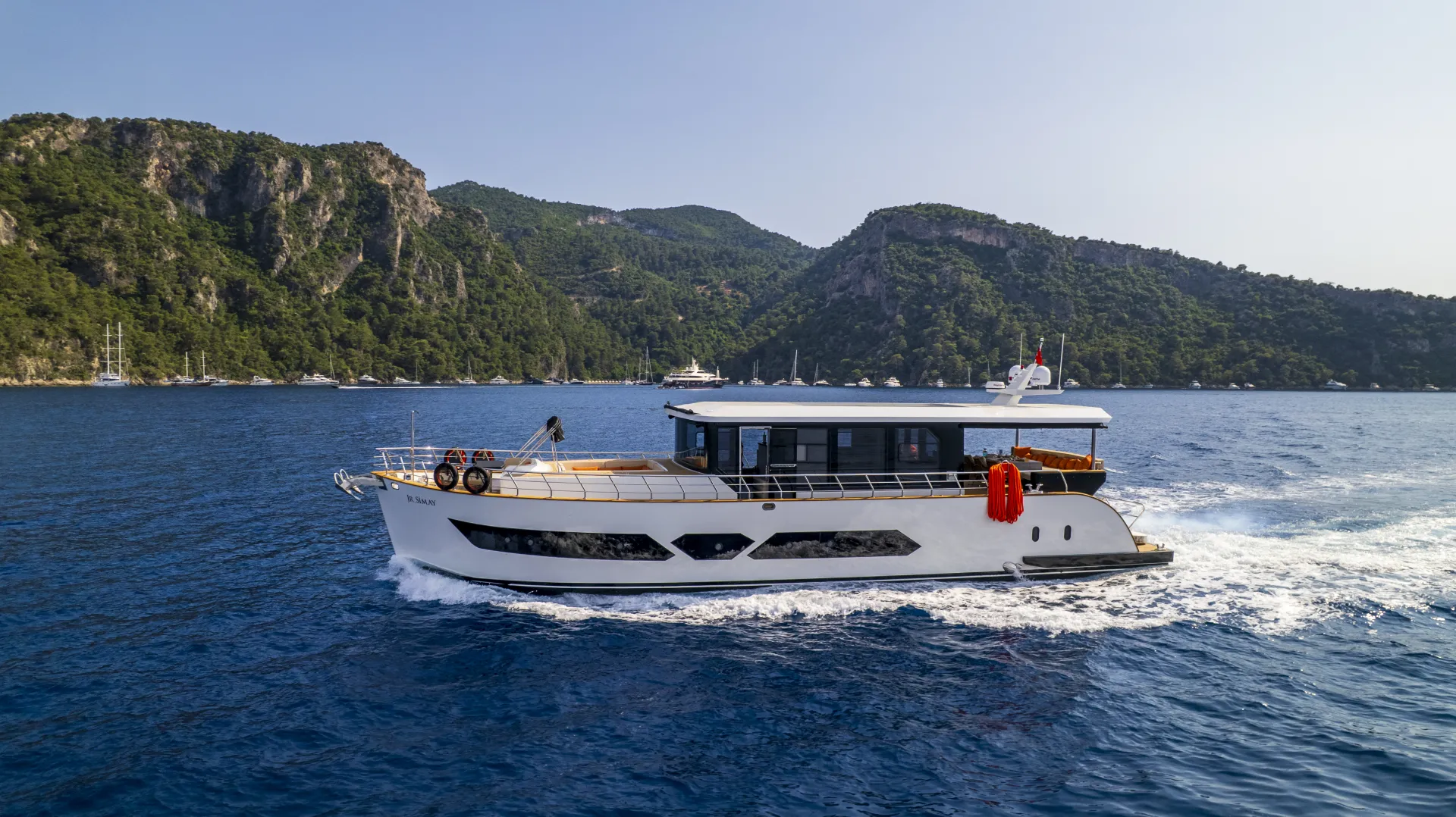 JUNIOR SIMAY A white motor yacht cruises through blue water with forested hills and rocky mountains in the background under a clear sky.