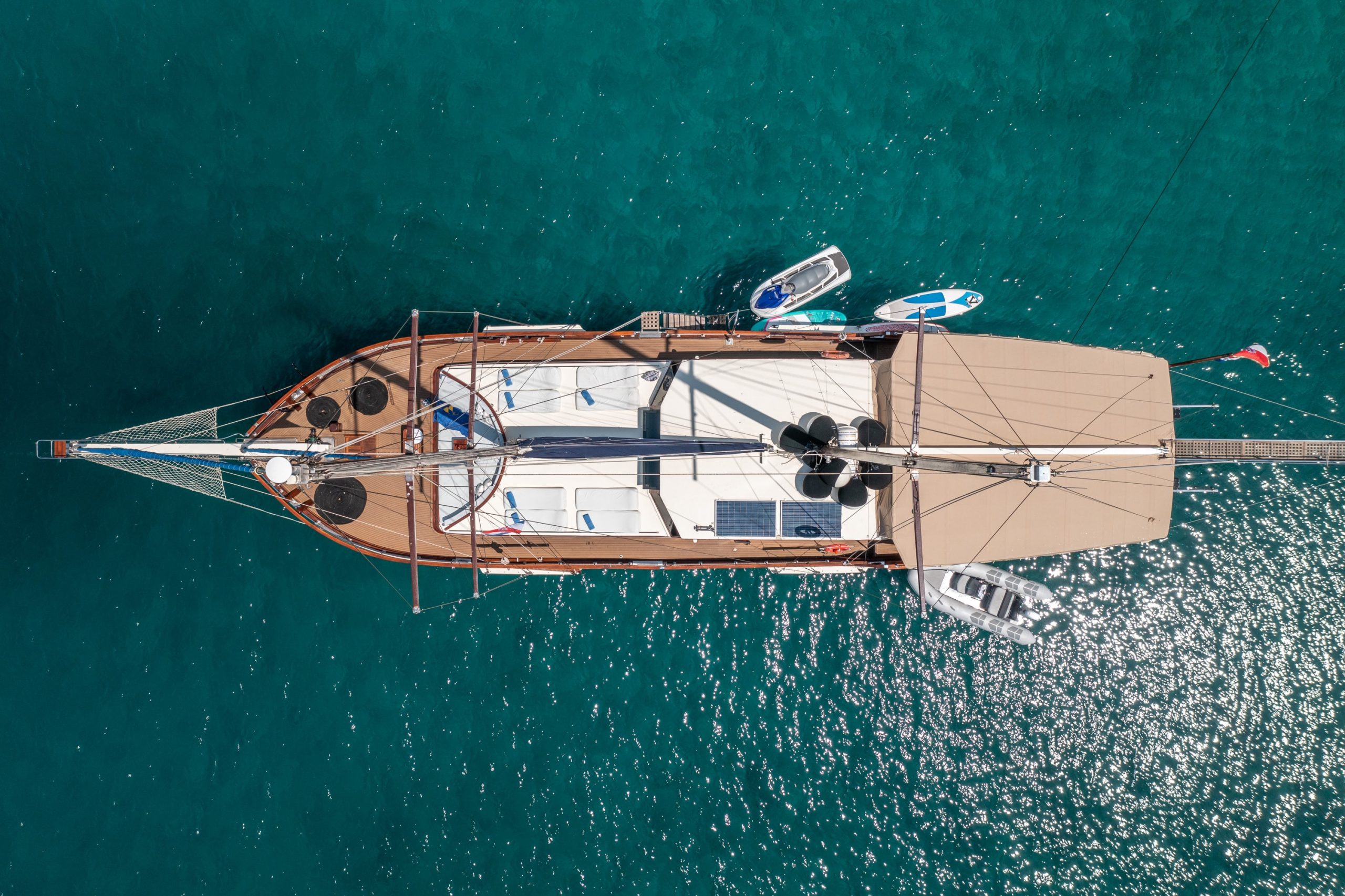 Aerial view of a large sailboat with a tan canopy floating on clear turquoise water, surrounded by small dinghies and paddleboards.