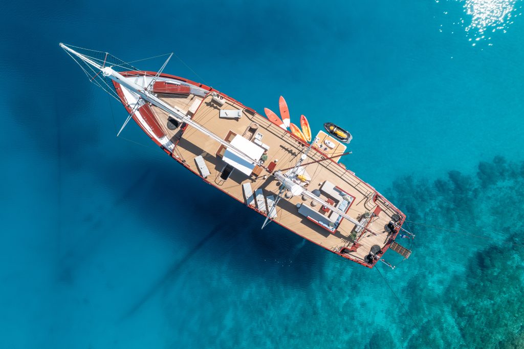ROMANCA Aerial view of a large sailboat with wooden decking floating on clear blue water, surrounded by kayaks and paddleboards, with shadows of the boat visible on the sea floor below.