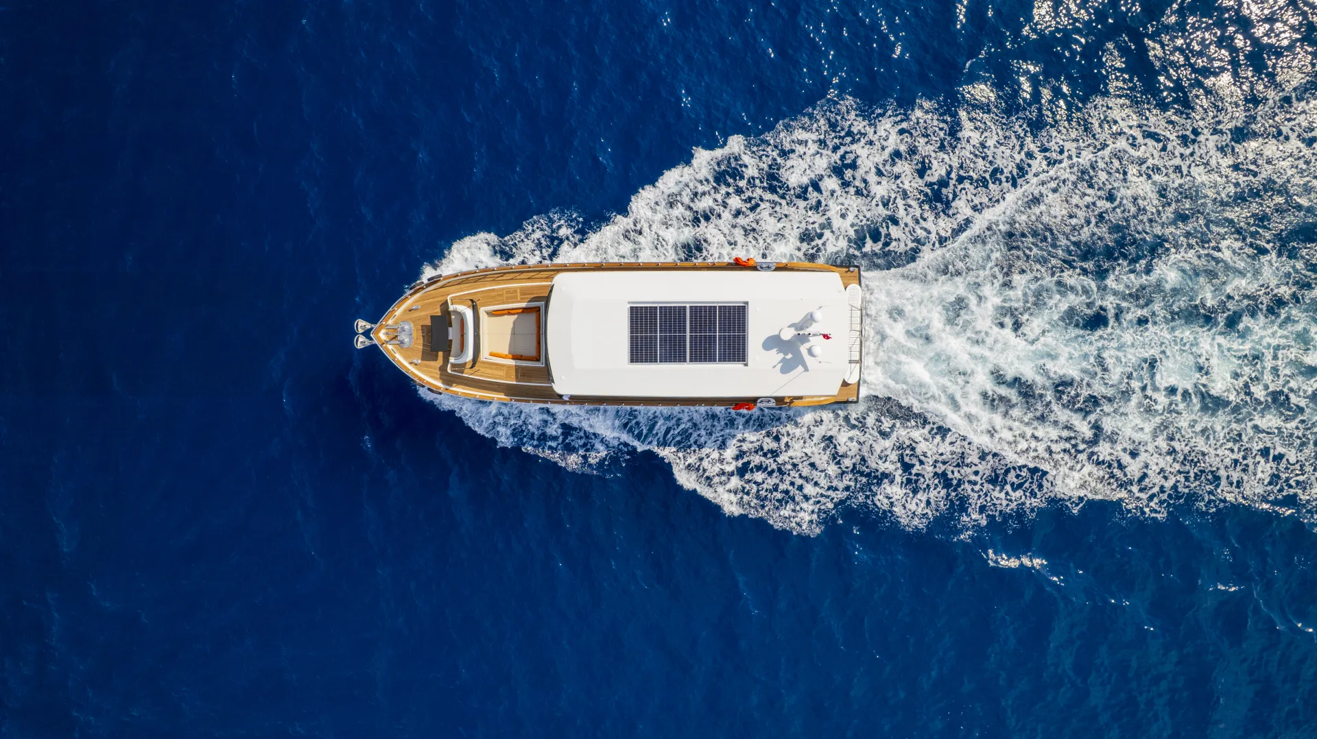 JUNIOR SIMAY Aerial view of a boat with a solar panel on its roof cruising through deep blue water, leaving a trail of white waves behind it.