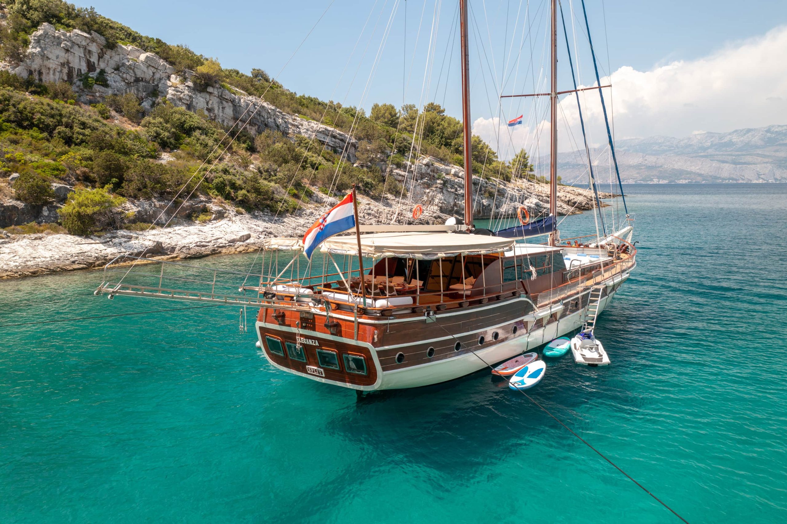 A wooden sailboat with a Croatian flag is anchored in clear turquoise water near a rocky, tree-covered coastline. Paddleboards float nearby, and mountains are visible in the distance under a partly cloudy sky.