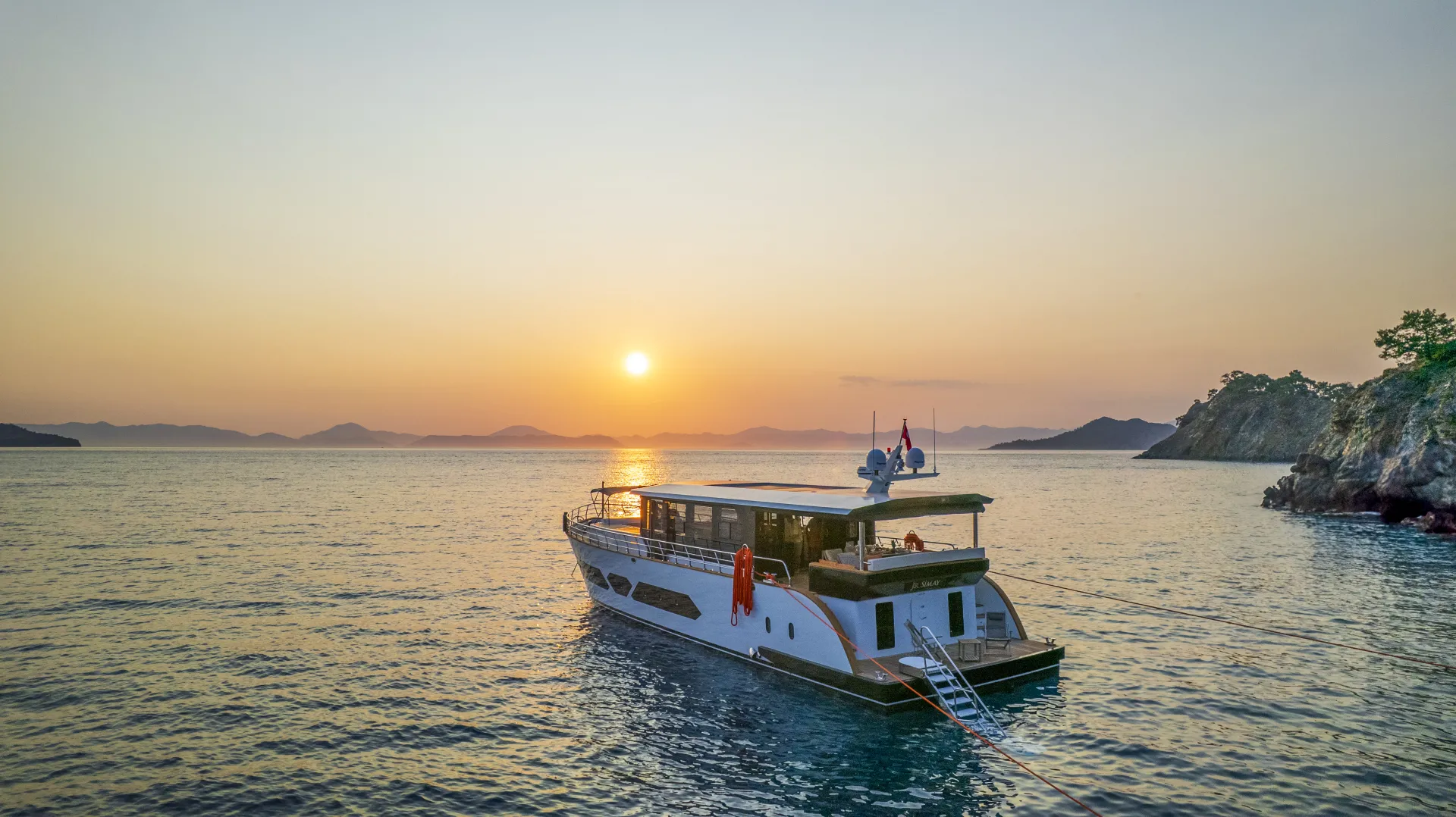 JUNIOR SIMAY A yacht anchored on calm water at sunset, with orange and yellow hues in the sky, distant mountains on the horizon, and rocky shorelines on either side.