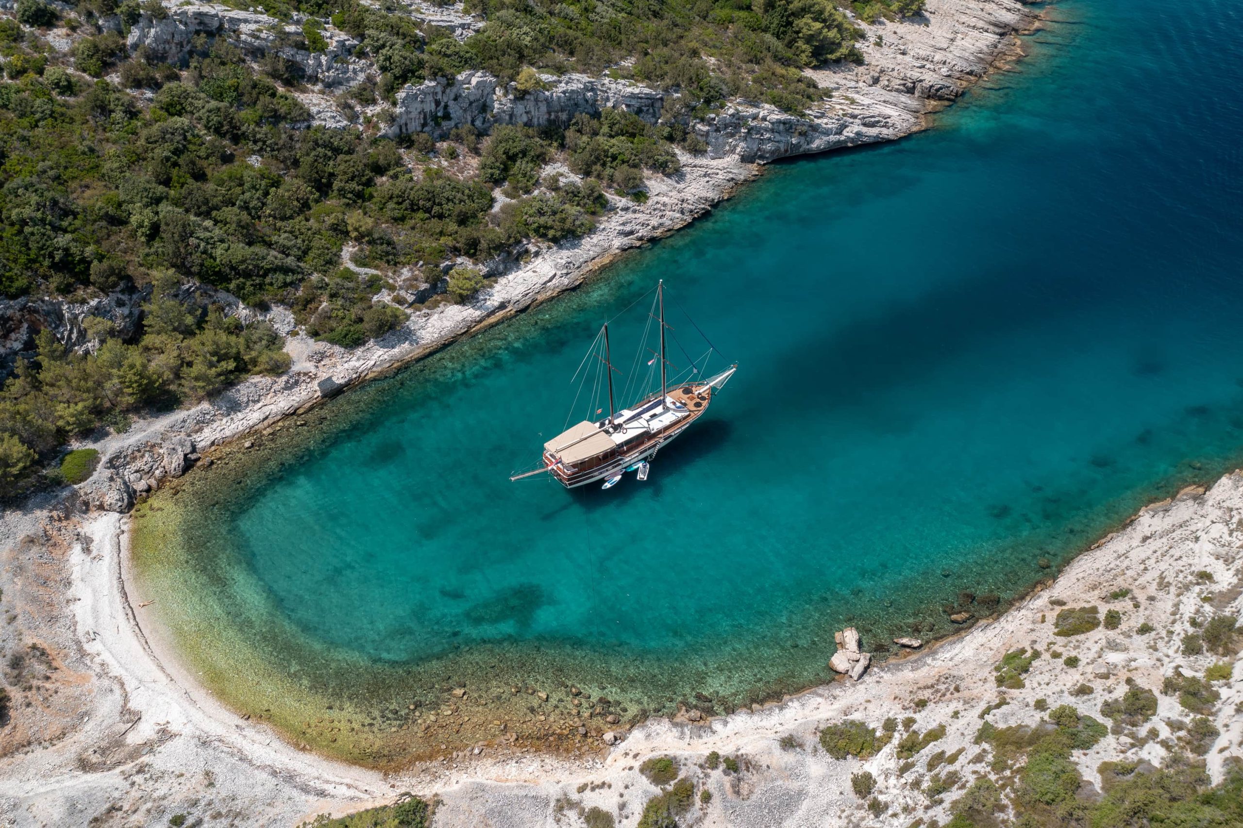 Aerial view of a sailboat anchored in a small, clear turquoise cove surrounded by rocky cliffs and greenery, with a narrow white-sand beach curving along the shoreline.