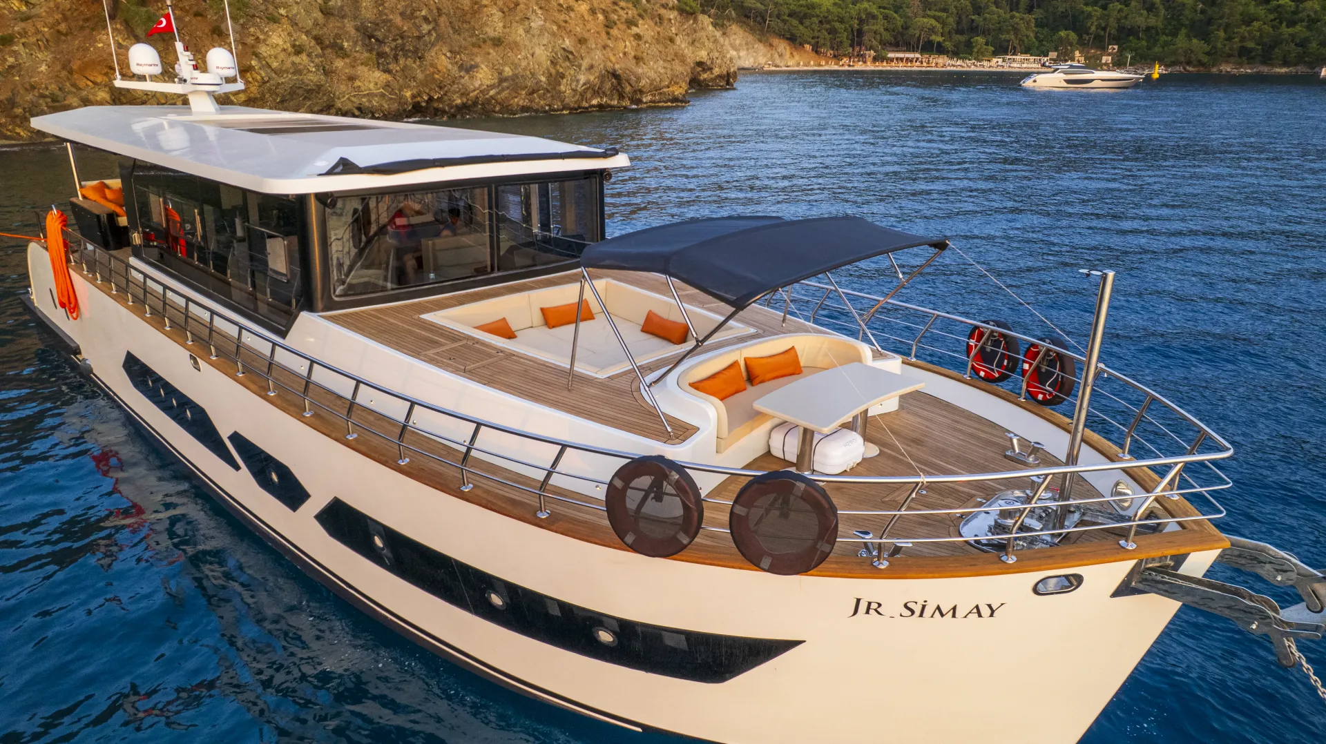 JUNIOR SIMAY A modern white yacht named JR SIMAY floats on calm blue water near a rocky coast, featuring sun loungers and a shaded seating area on its deck.