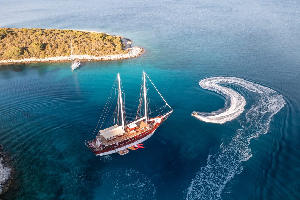 ROMANCA Aerial view of a large sailboat anchored in clear blue water near a green, rocky coastline, with a smaller boat making a circular wake nearby. Another sailboat is moored close to the shore.
