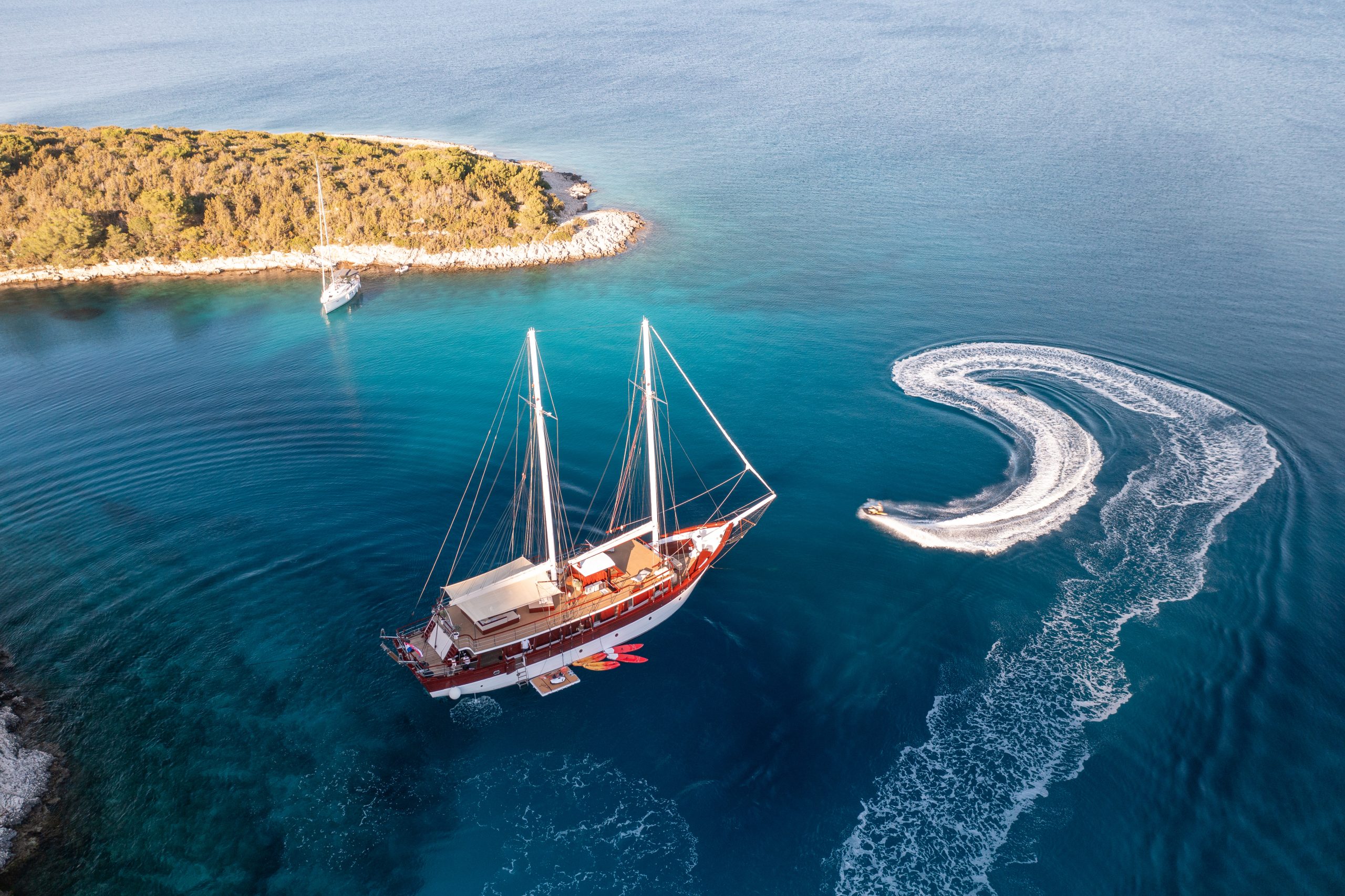 ROMANCA Aerial view of a large sailboat anchored near a small island with lush greenery, while a speedboat circles in the turquoise water, creating white trails behind it. Another sailboat is near the coast.