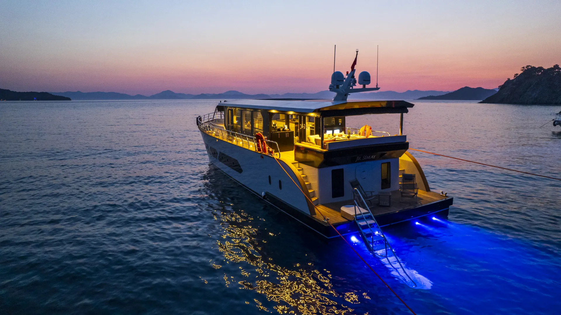 JUNIOR SIMAY A luxury yacht with illuminated blue underwater lights is anchored on calm water at sunset, with gentle waves and distant mountains silhouetted against a colorful sky.