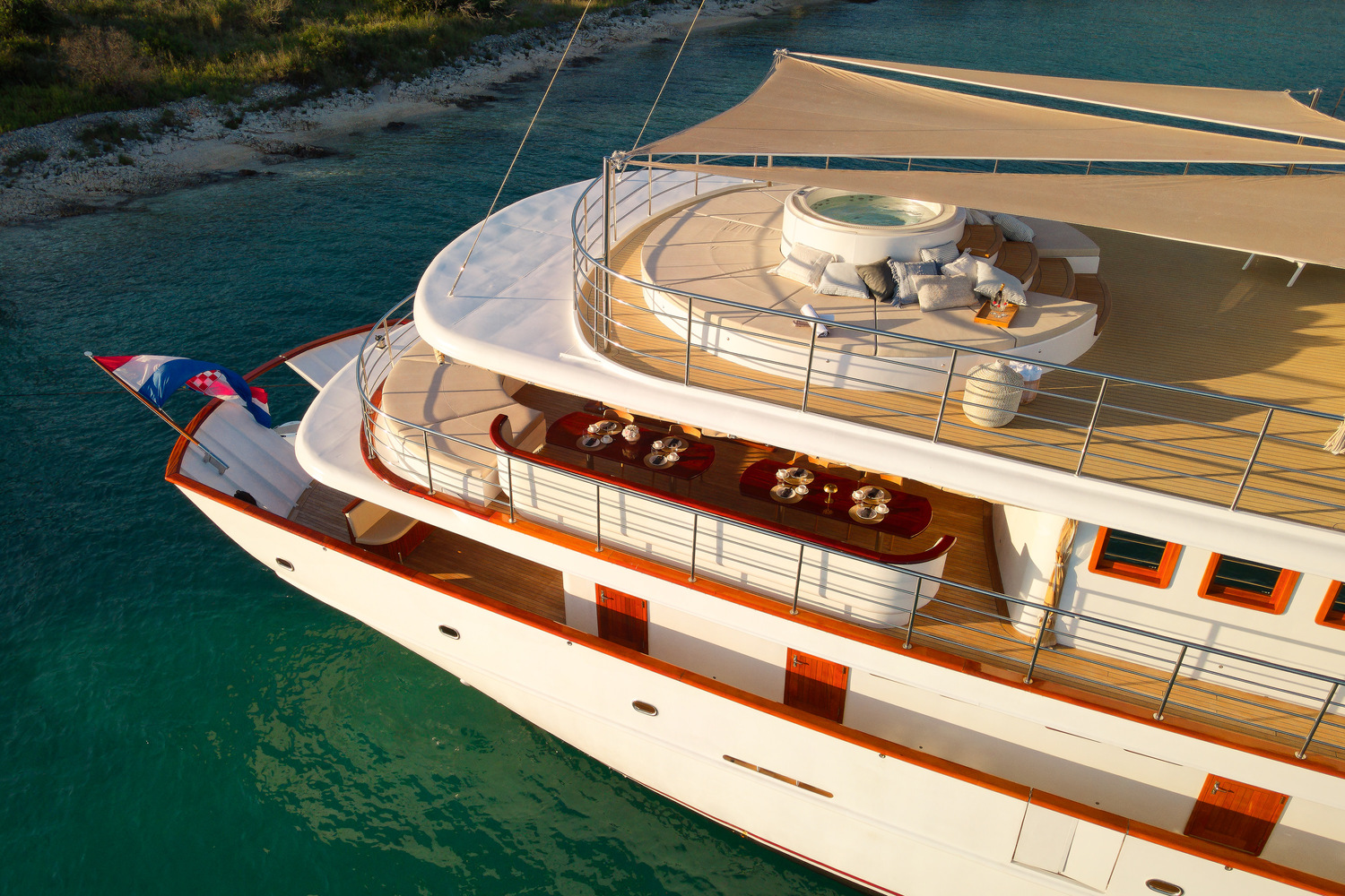 A luxurious white yacht anchored in clear turquoise water, featuring an upper deck with a round jacuzzi, sun loungers, a shaded area, and an elegantly set dining table below. One person lounges near the jacuzzi.