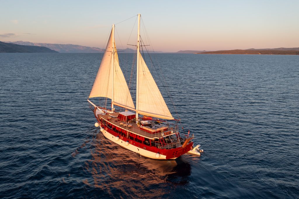 ROMANCA A red and white sailboat with two large sails glides on calm blue water at sunset, with distant mountains and coastline in the background.