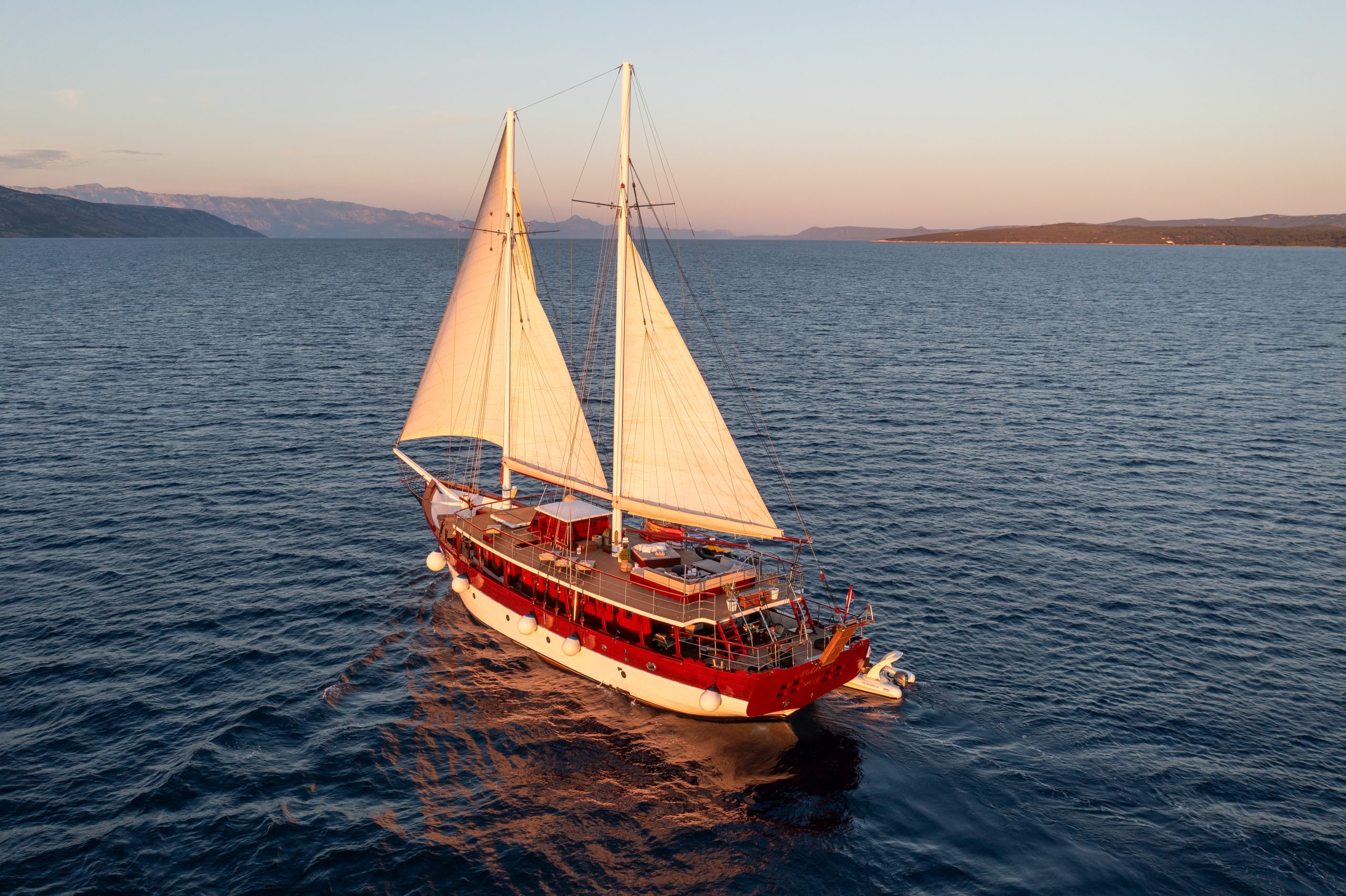 ROMANCA A red and white sailboat with two large sails glides across calm blue water during sunset, with distant land visible on the horizon under a clear sky.