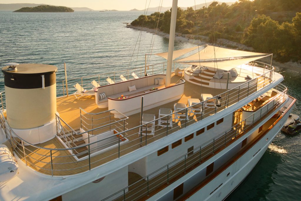 A luxury yacht with a spacious upper deck featuring lounge chairs, a shaded seating area, and a hot tub, anchored near a forested coastline at sunset.