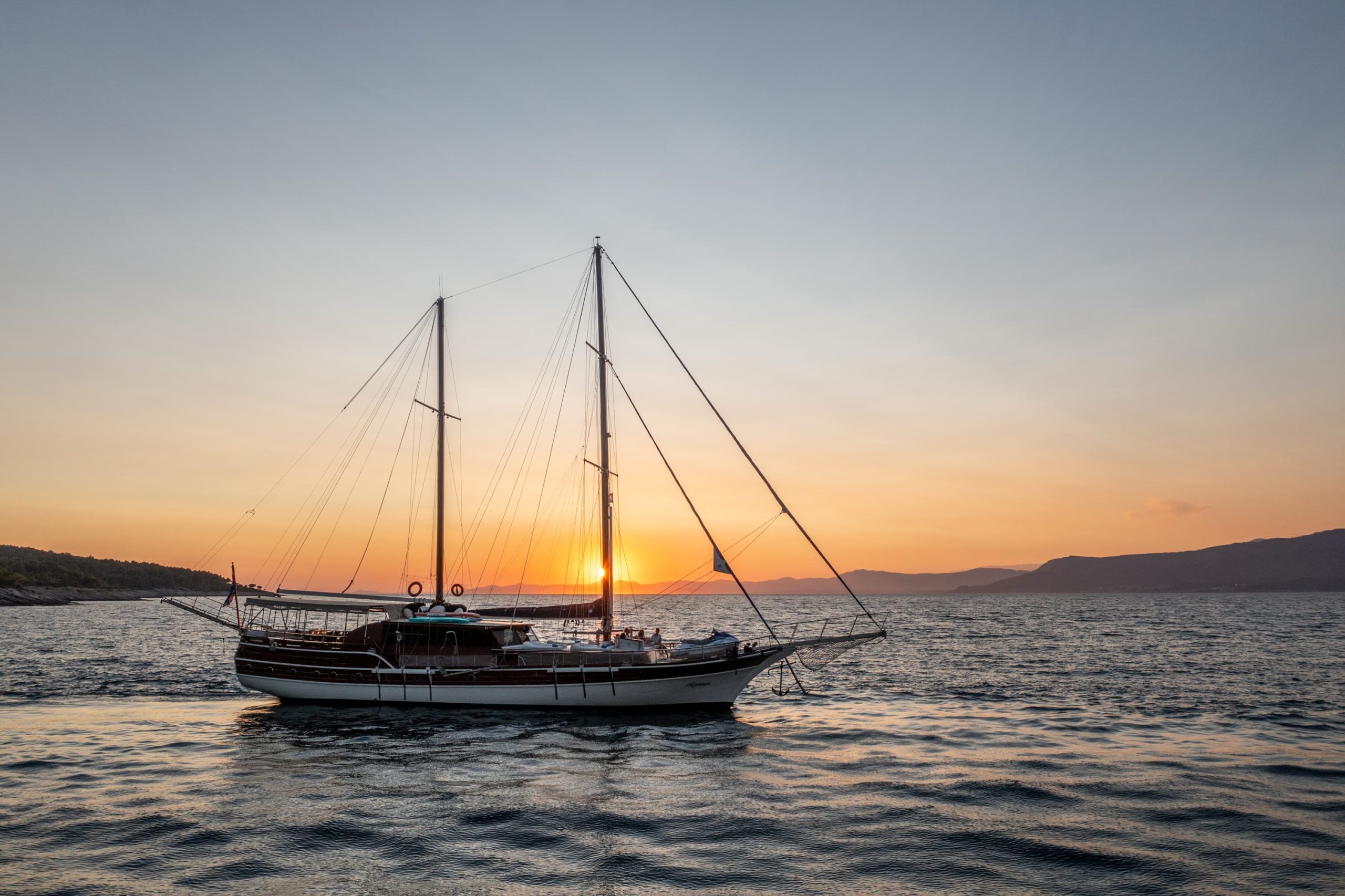 A sailboat with two masts floats on calm water at sunset, with the sun partially visible near the horizon and distant hills in the background. The sky is clear, transitioning from orange to blue.