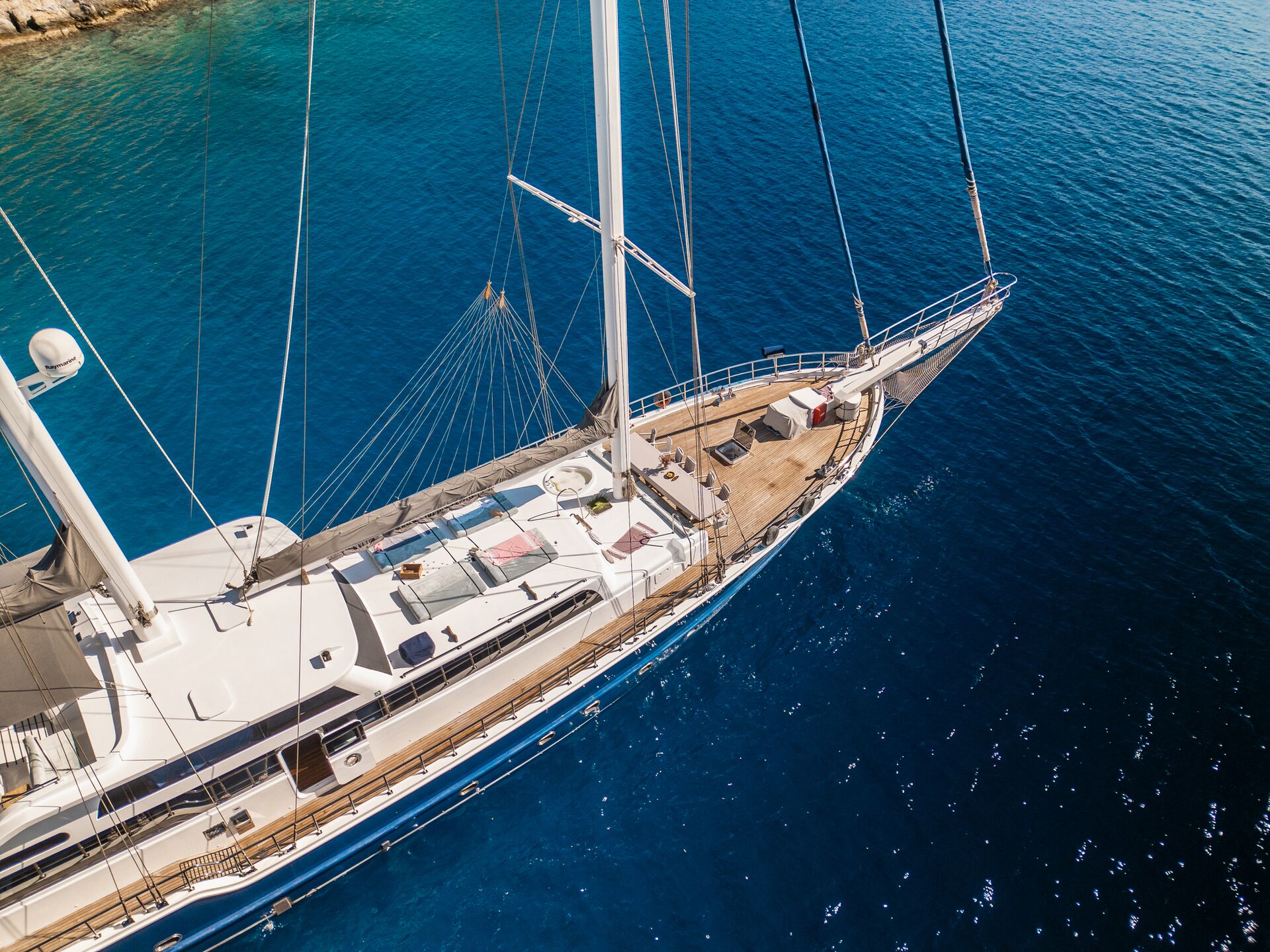 Aerial view of a luxury sailboat with a wooden deck and sunbathing area, anchored in clear, deep blue water near a rocky coastline.