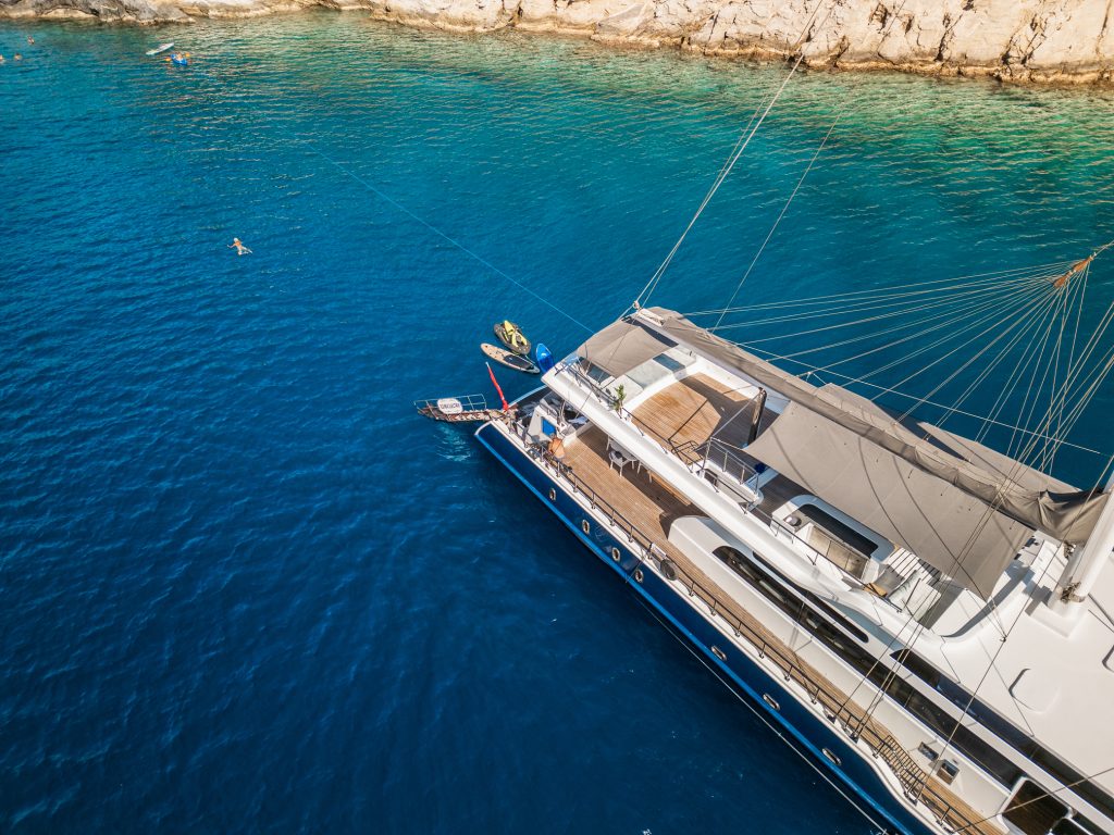 A large yacht is anchored in clear blue water near a rocky coastline. A few people are swimming in the sea, and a small boat is docked next to the yacht. The sun is shining, and the water is calm.