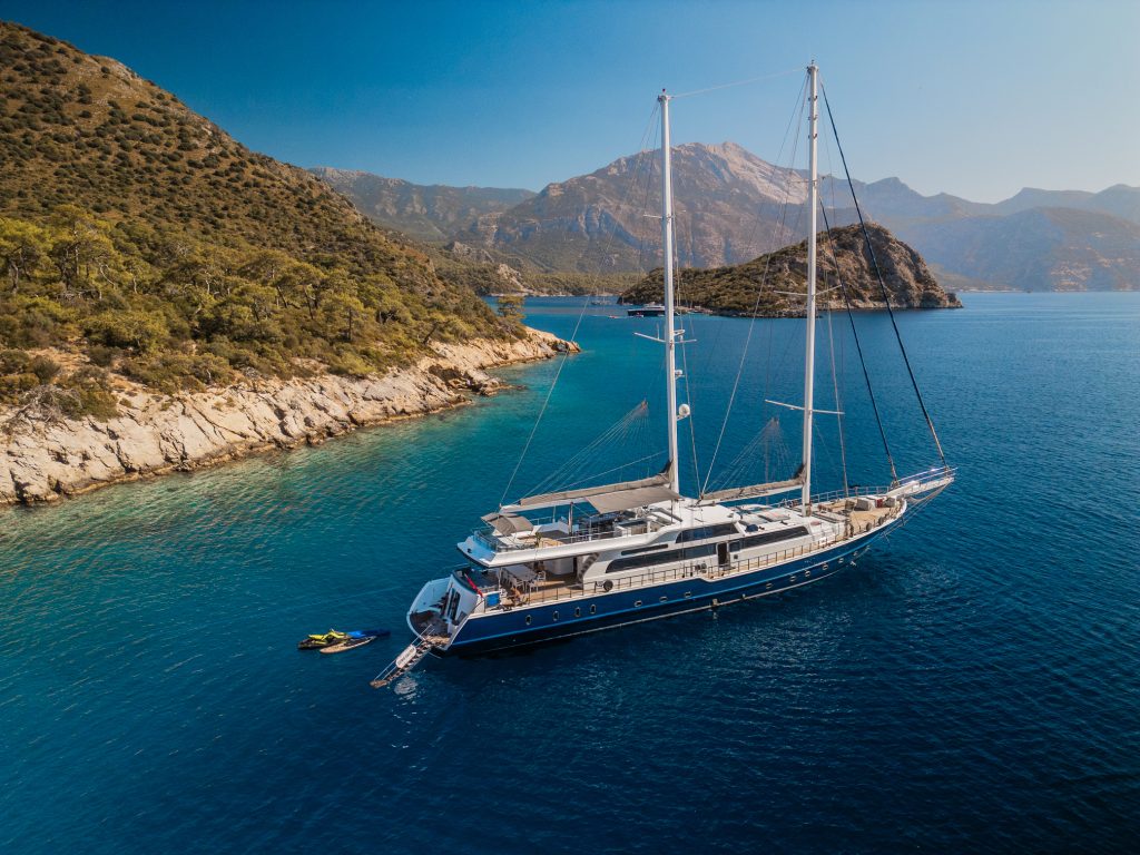 A large sailboat yacht anchored in clear blue water near a rocky, green coastline with mountains in the background under a clear sky. A small boat and jet ski are next to the yacht.
