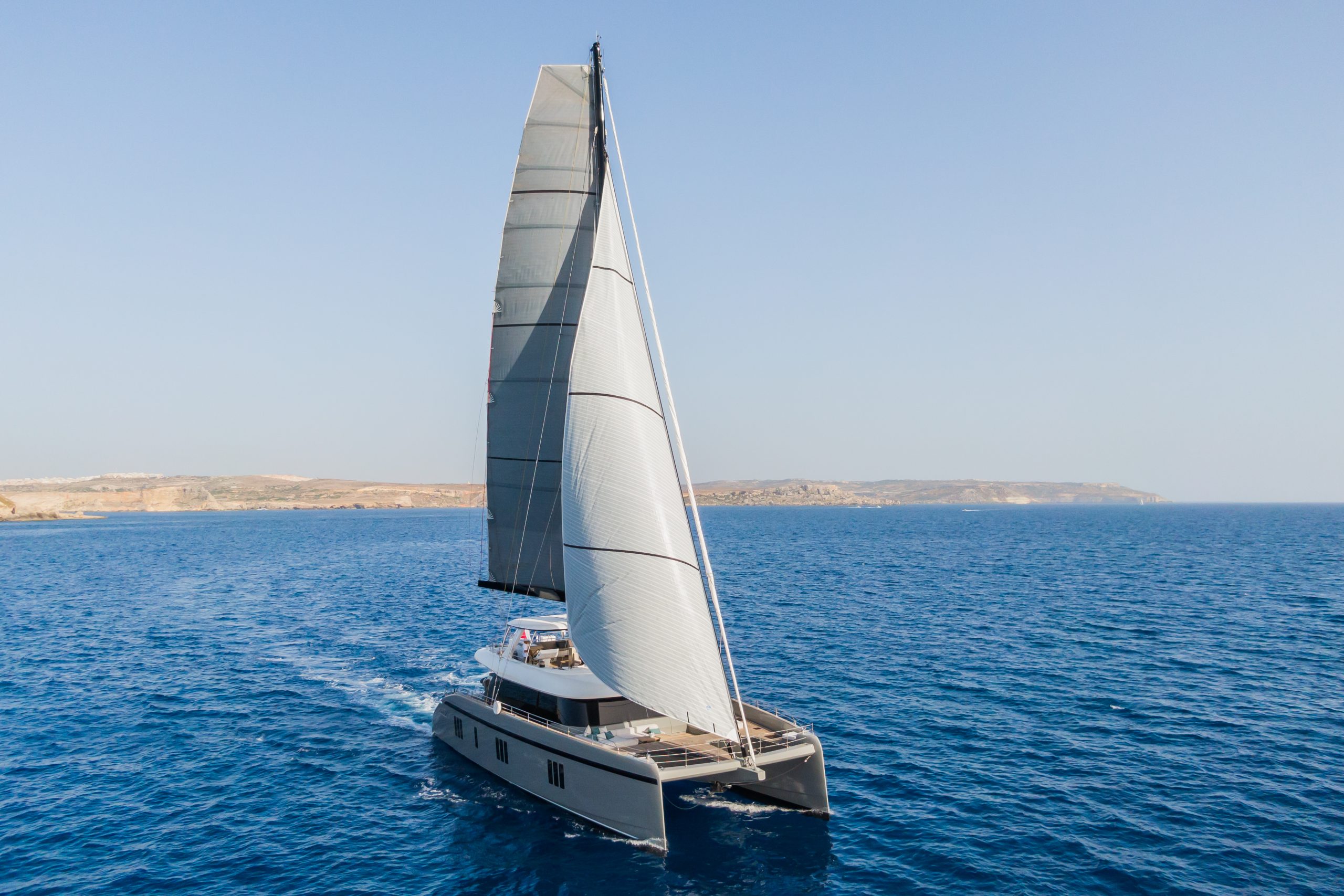 GREYB A modern catamaran yacht with large sails glides across deep blue ocean water under a clear sky, with distant land visible on the horizon.