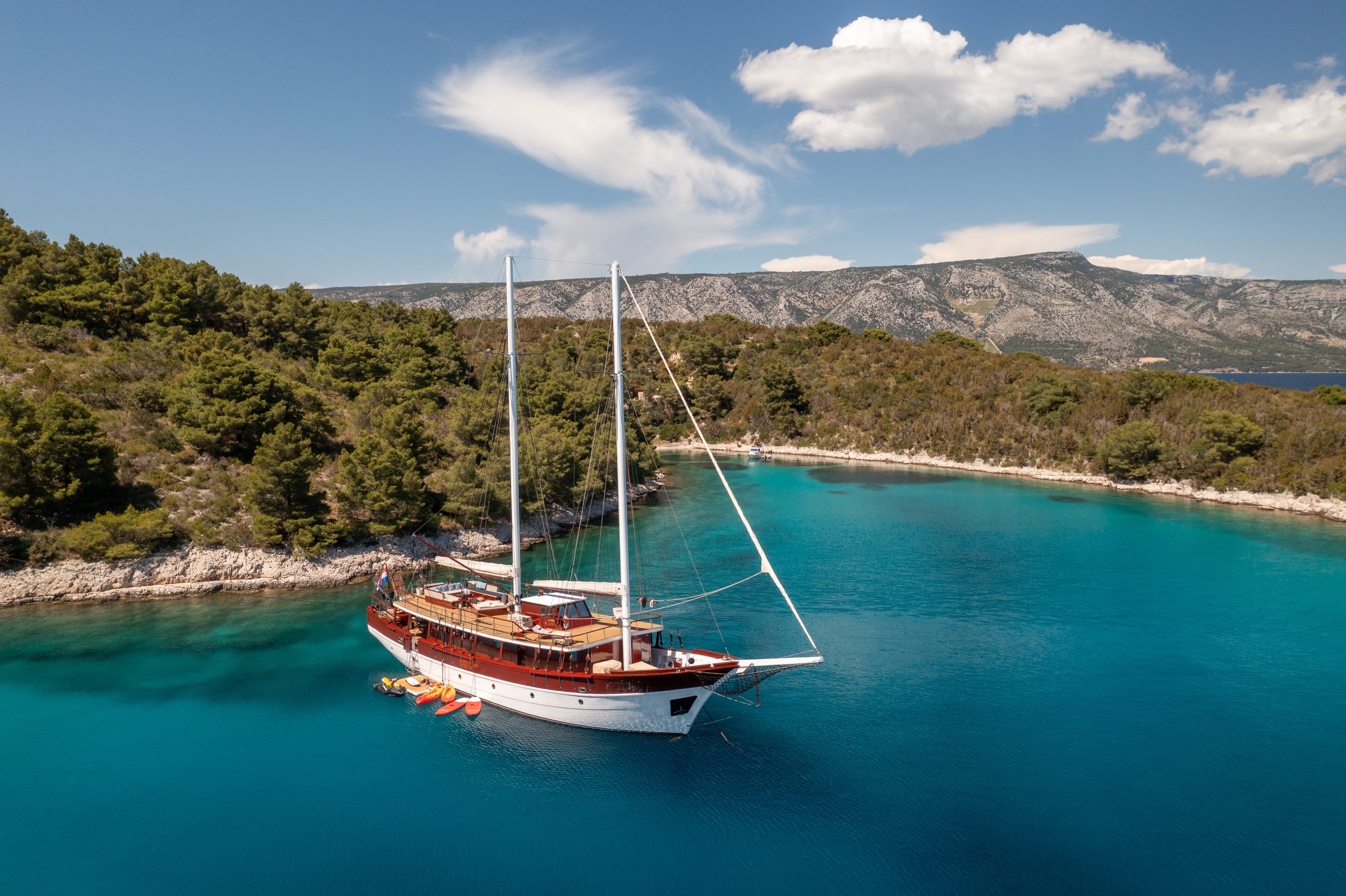 ROMANCA A wooden sailboat is anchored in clear turquoise water near a forested coastline with rocky shores. Paddleboards float beside the boat. Green hills and a mountain range are visible under a blue sky with scattered clouds.
