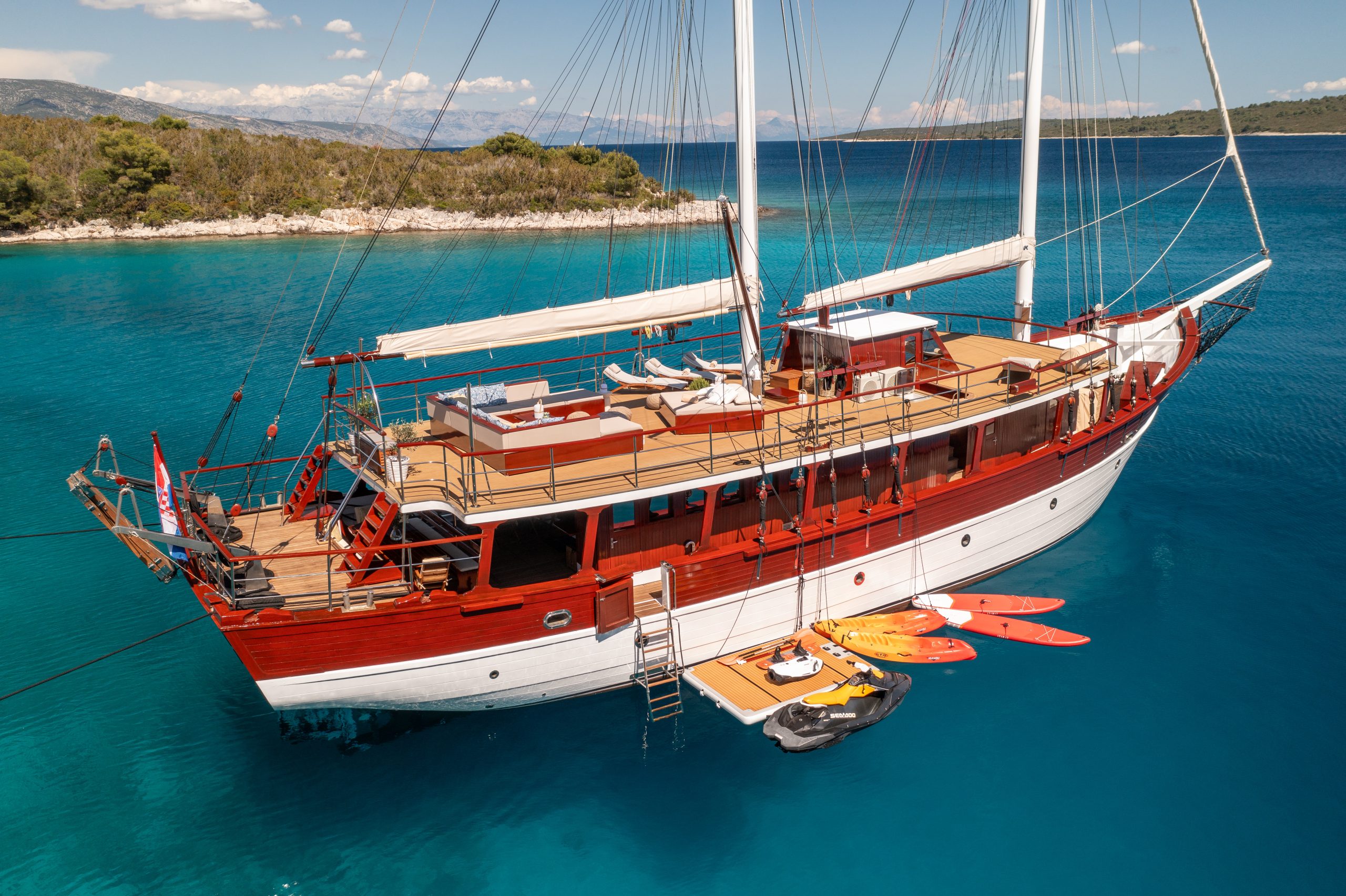 ROMANCA A large wooden sailing yacht with white and red accents is anchored in clear blue water near a rocky, tree-lined shore. Several kayaks and a small boat float beside it, and the deck is set up for relaxation.