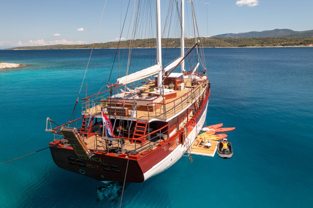 ROMANCA A large wooden sailboat with sun loungers on deck is anchored in clear turquoise water near a rocky shore. Paddleboards, kayaks, and a small inflatable boat float nearby. Distant hills and a blue sky complete the scene.