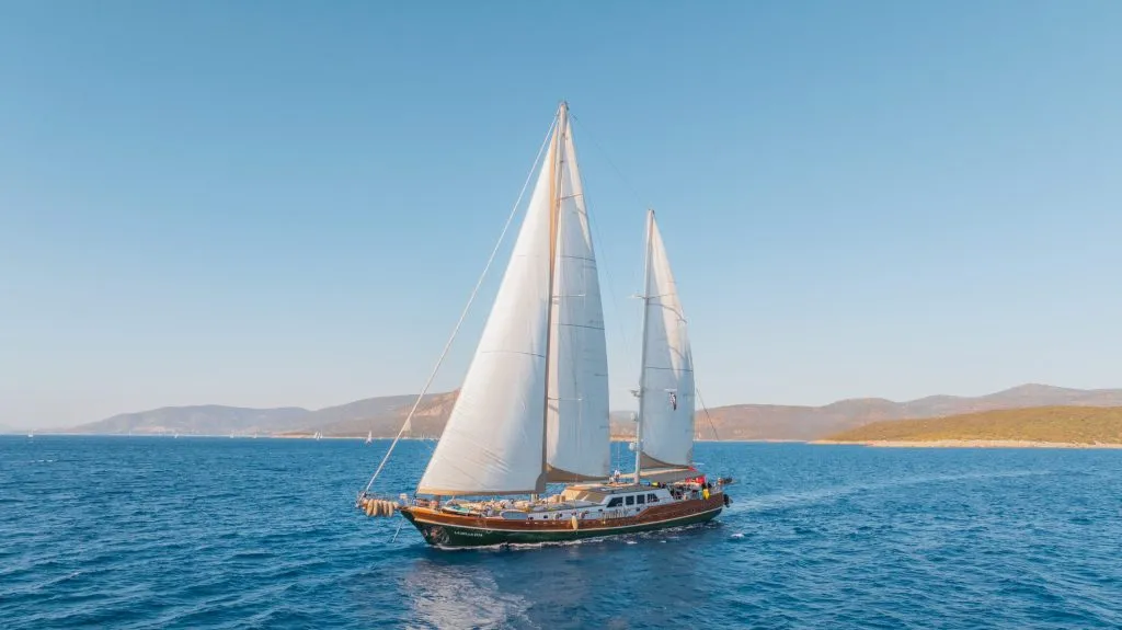 A large sailboat with two white sails glides across a calm blue sea under a clear sky, with distant hills and coastline visible in the background.