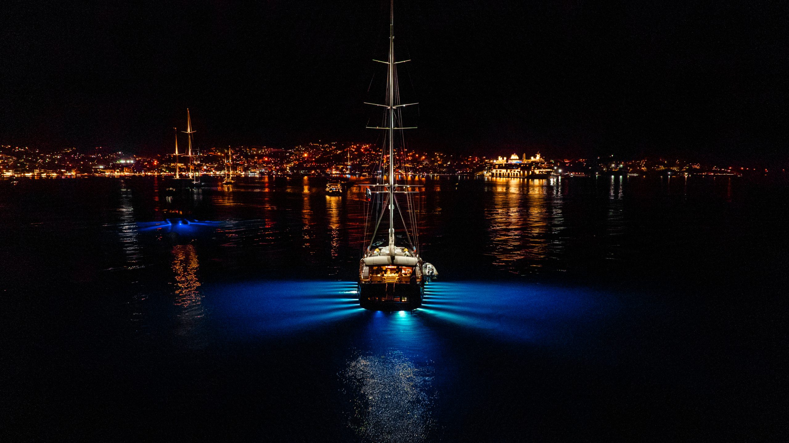 A sailboat illuminated by blue underwater lights floats on calm water at night, with city lights and buildings glowing along the distant shoreline.