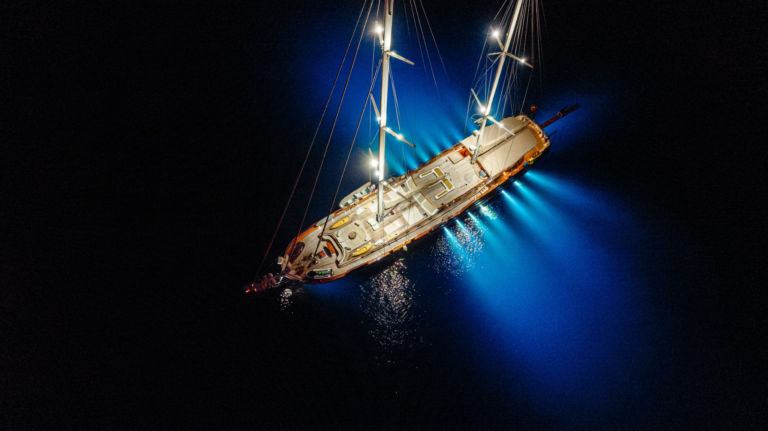 A sailboat illuminated by bright white and blue lights floats on dark water at night, casting a dramatic glow and reflections around its hull.