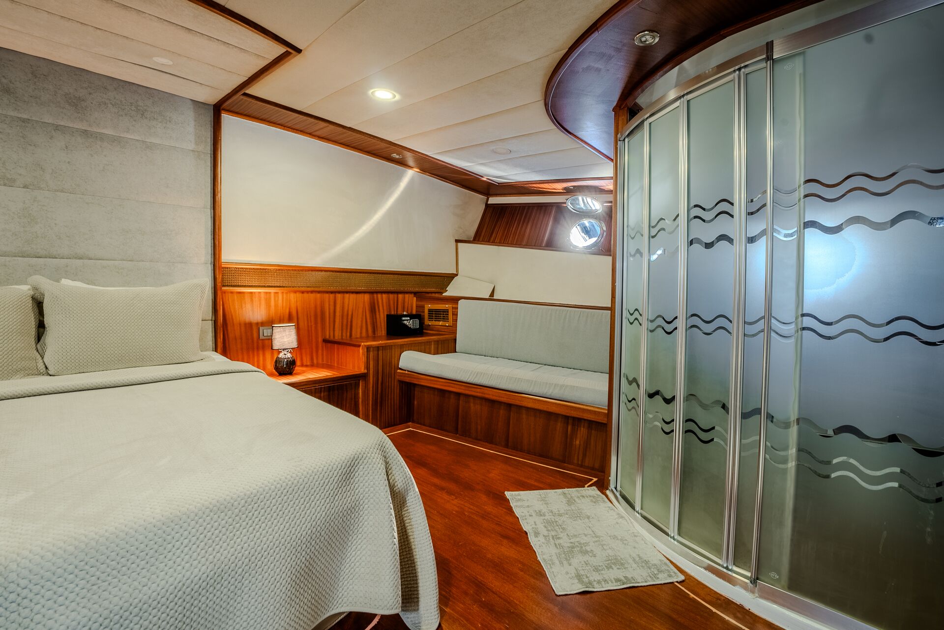 A cozy yacht cabin with a single bed, cushioned bench, wood paneling, a small nightstand, and a curved glass shower with frosted wave patterns. Warm lighting and modern decor create an inviting atmosphere.