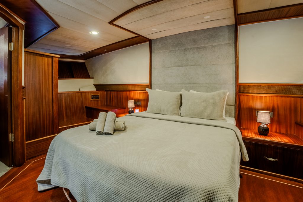 A cozy bedroom with a neatly made bed, beige bedding, and two rolled towels on top. The room features warm wooden paneling, soft lighting, and two small lamps on bedside tables.