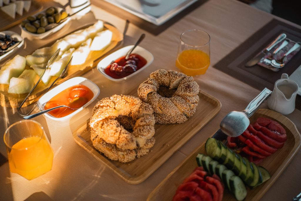 A breakfast table with two sesame bagels, sliced cucumbers and tomatoes, fruit, olives, sauces, two glasses of orange juice, and utensils arranged neatly on a beige tablecloth.