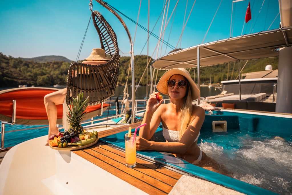 A woman in a sunhat and sunglasses relaxes in a yachts hot tub, holding a drink with straws. A fruit platter sits nearby, and another person lounges in a swinging chair. The scene is sunny with water and greenery in the background.