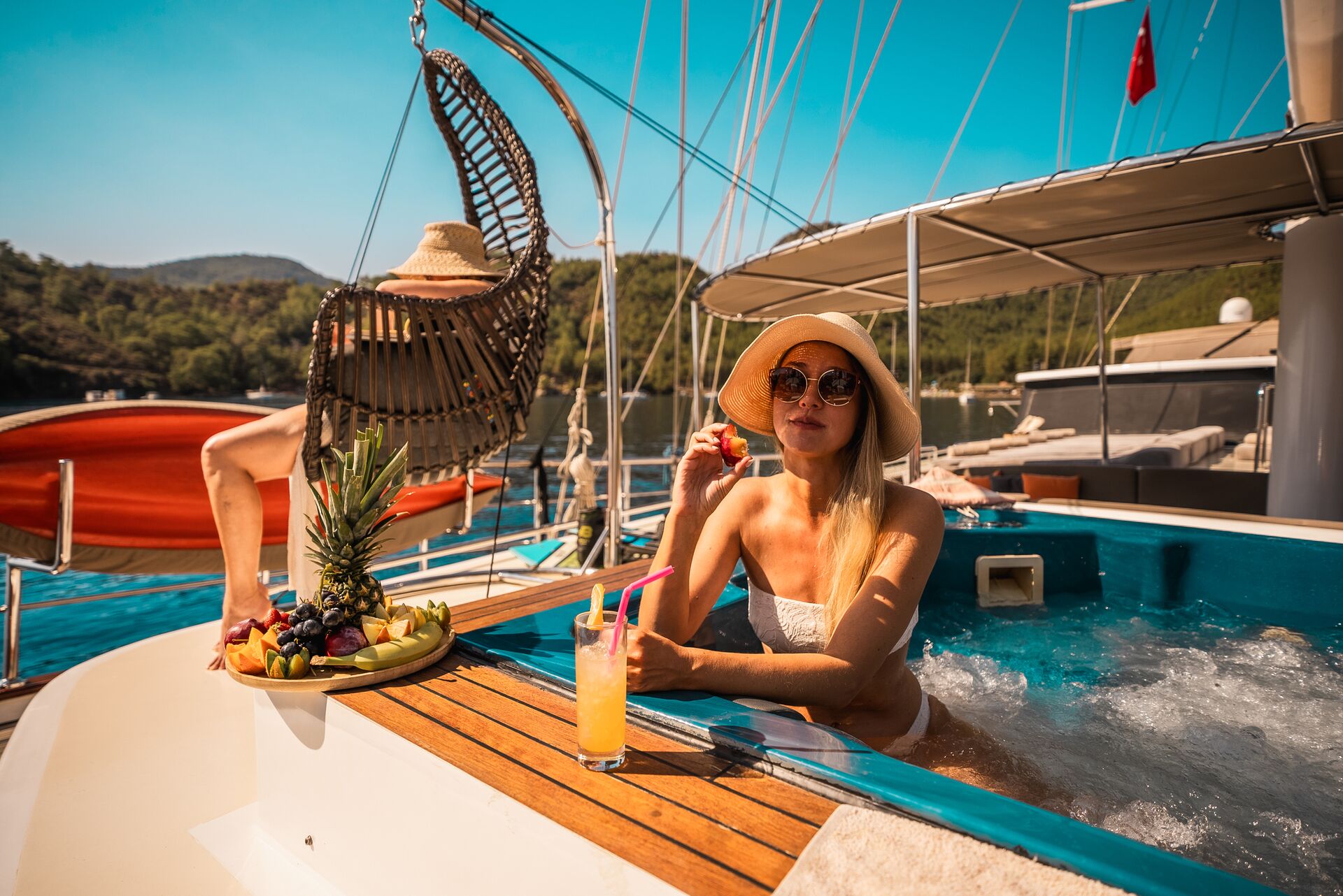 A woman in a sunhat and sunglasses relaxes in a yachts hot tub, holding a drink with straws. A fruit platter sits nearby, and another person lounges in a swinging chair. The scene is sunny with water and greenery in the background.