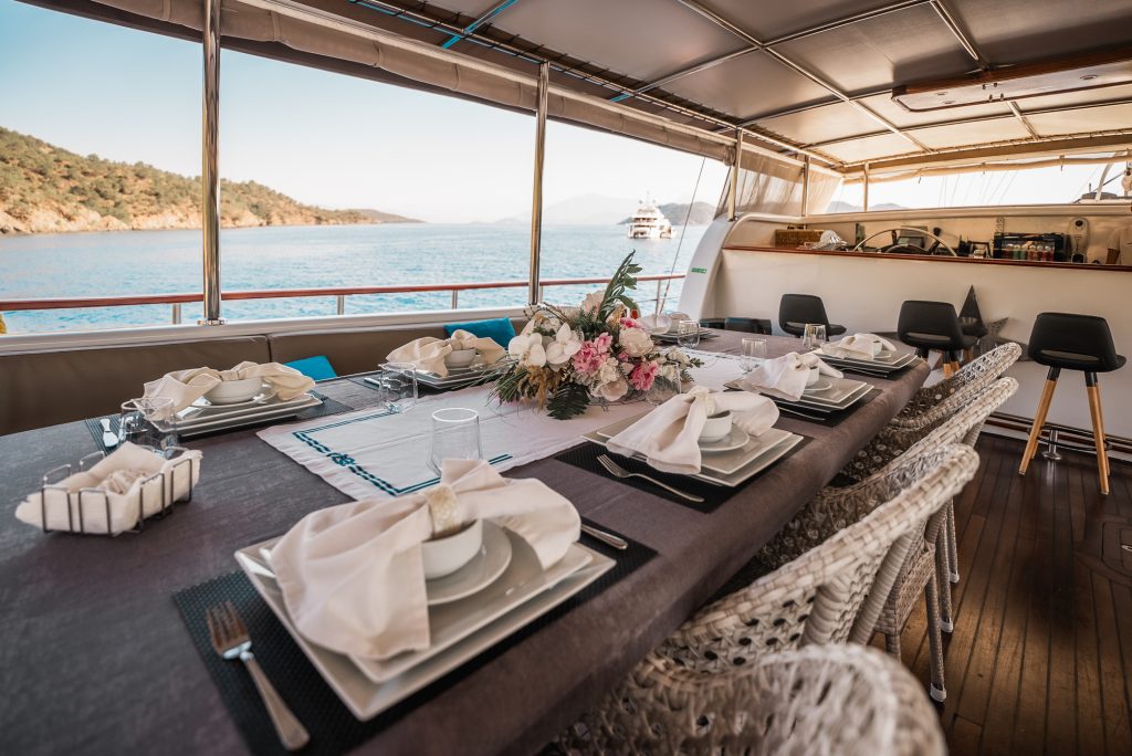 A dining table set for six with elegant plates, napkins, and a floral centerpiece on a yacht deck, overlooking calm blue water and distant boats, with coastal hills in the background.