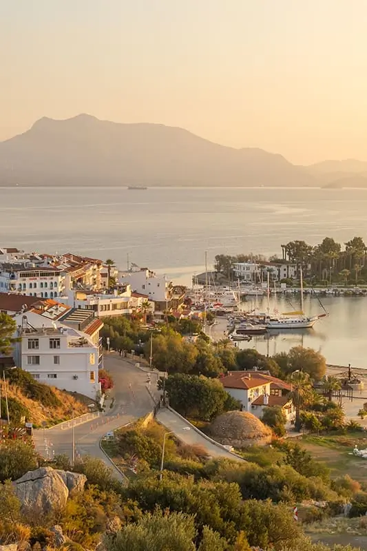 Coastal town at sunset with boats docked in a marina, white buildings, winding roads, and mountains in the background, all bathed in a soft golden light.