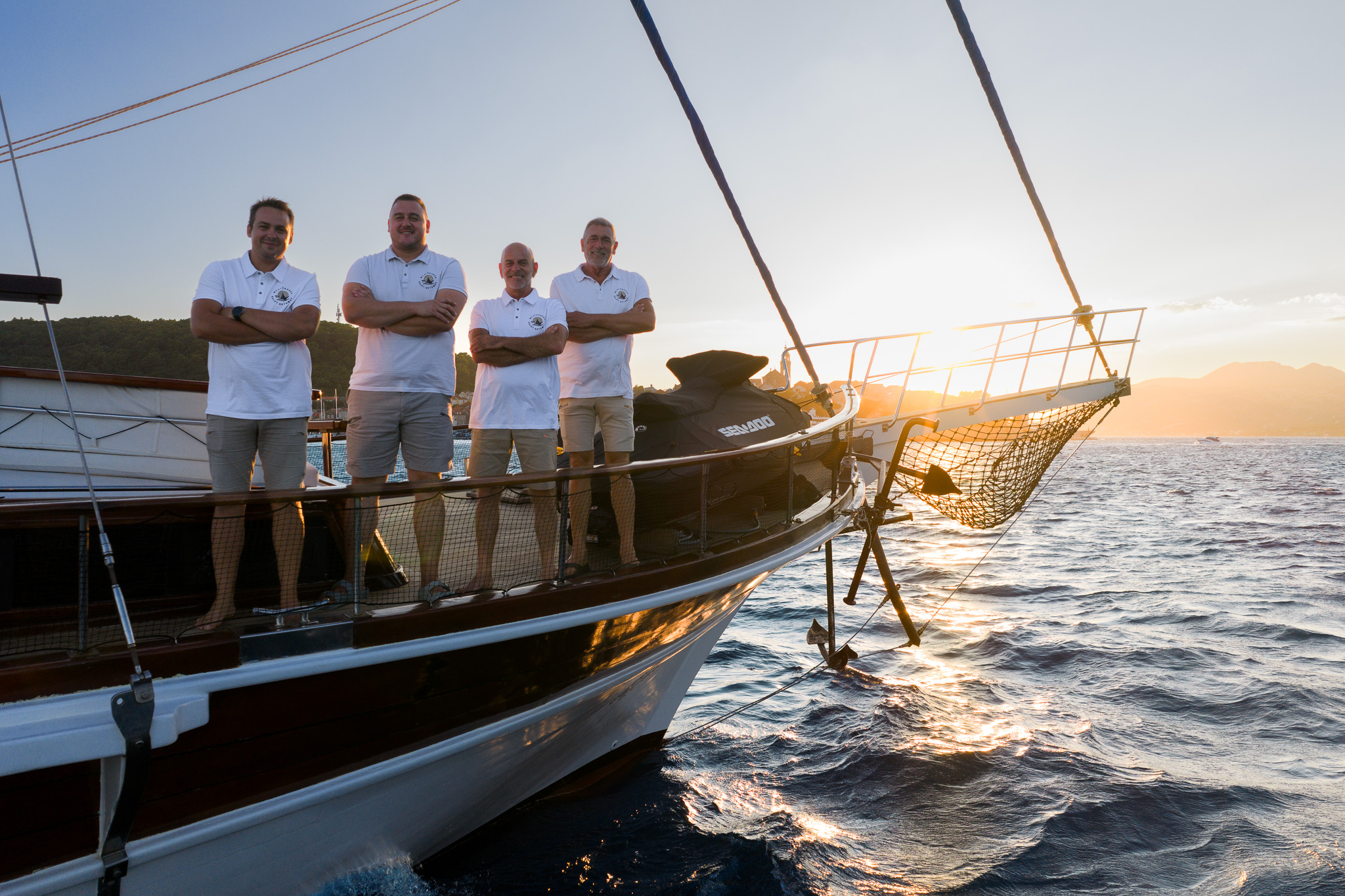 Four men in white polo shirts and shorts stand on the deck of a boat at sunset, with arms crossed, overlooking the sea and distant mountains. The water is slightly choppy and the sky is clear.